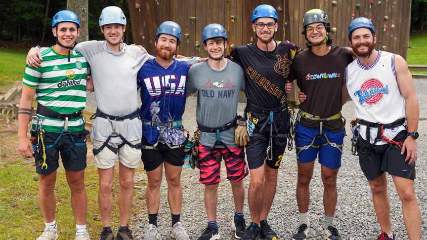 Seven smiling camp counselors gathered near the rock climbing tower, emphasizing the camaraderie and joy of summer jobs in the great outdoors