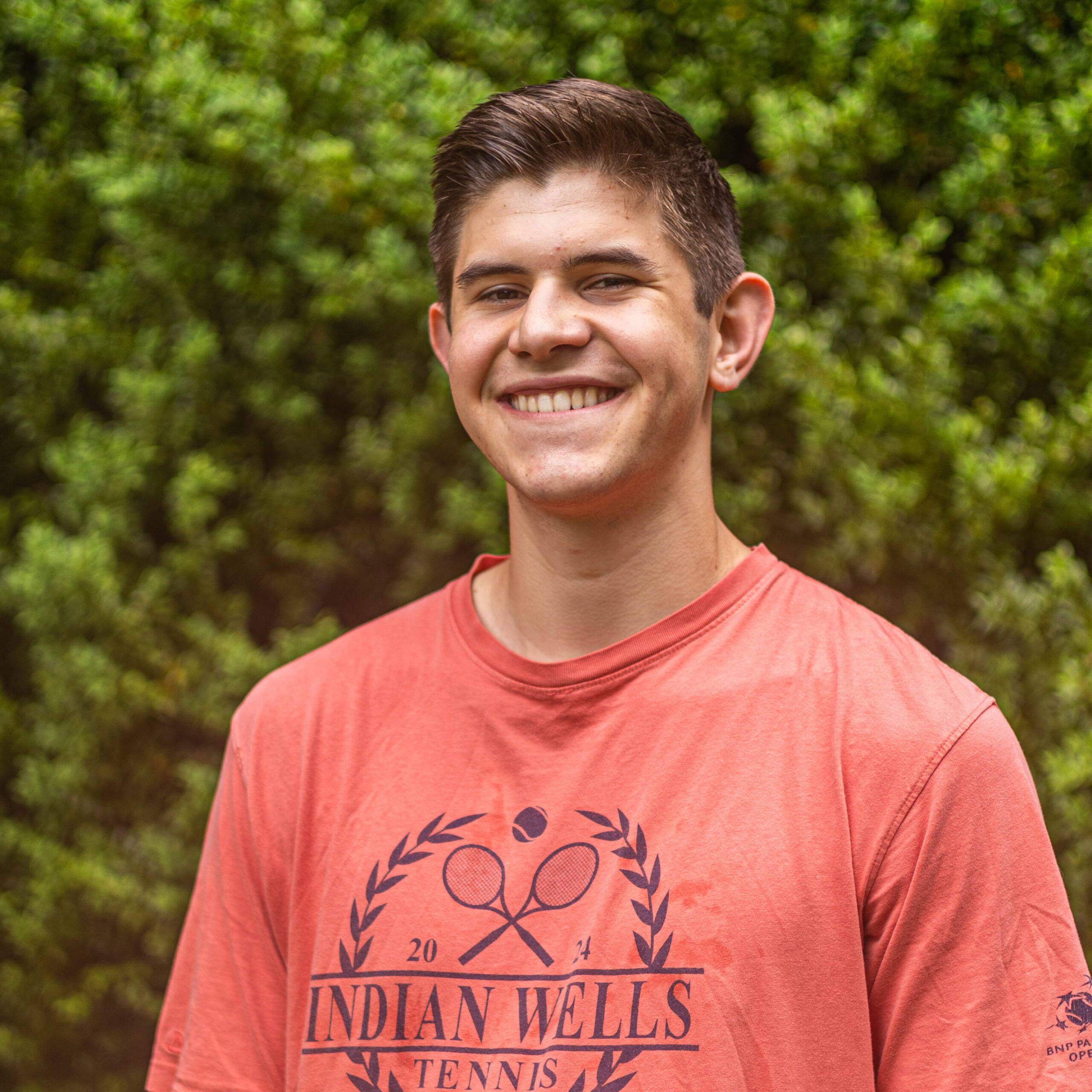Smiling man wearing a coral Indiana Wells Tennis t-shirt outdoors with green foliage in the background.