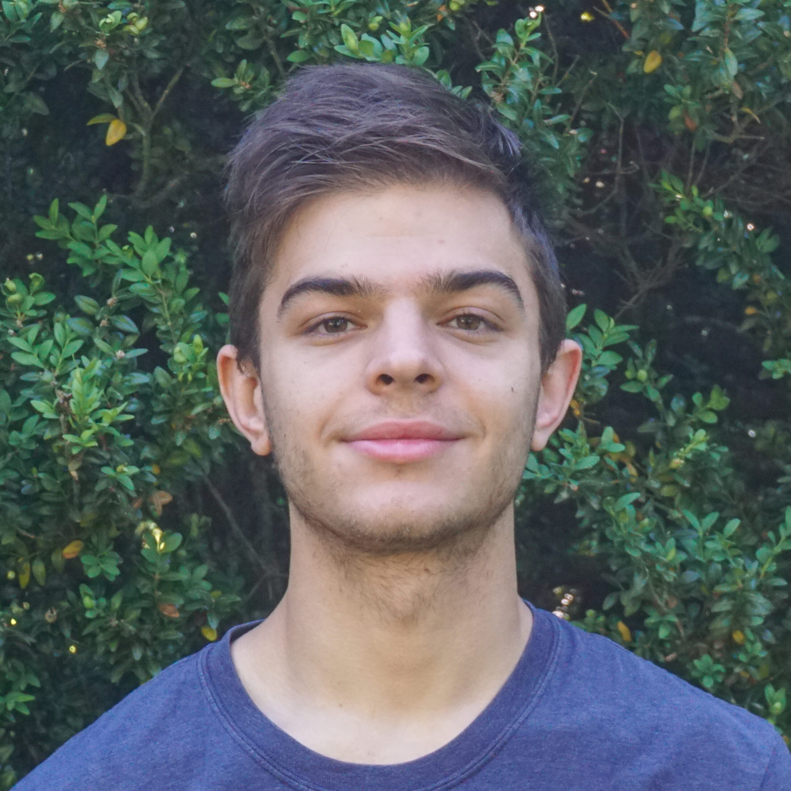 Close-up of a young man outdoors with green leafy bushes in the background.