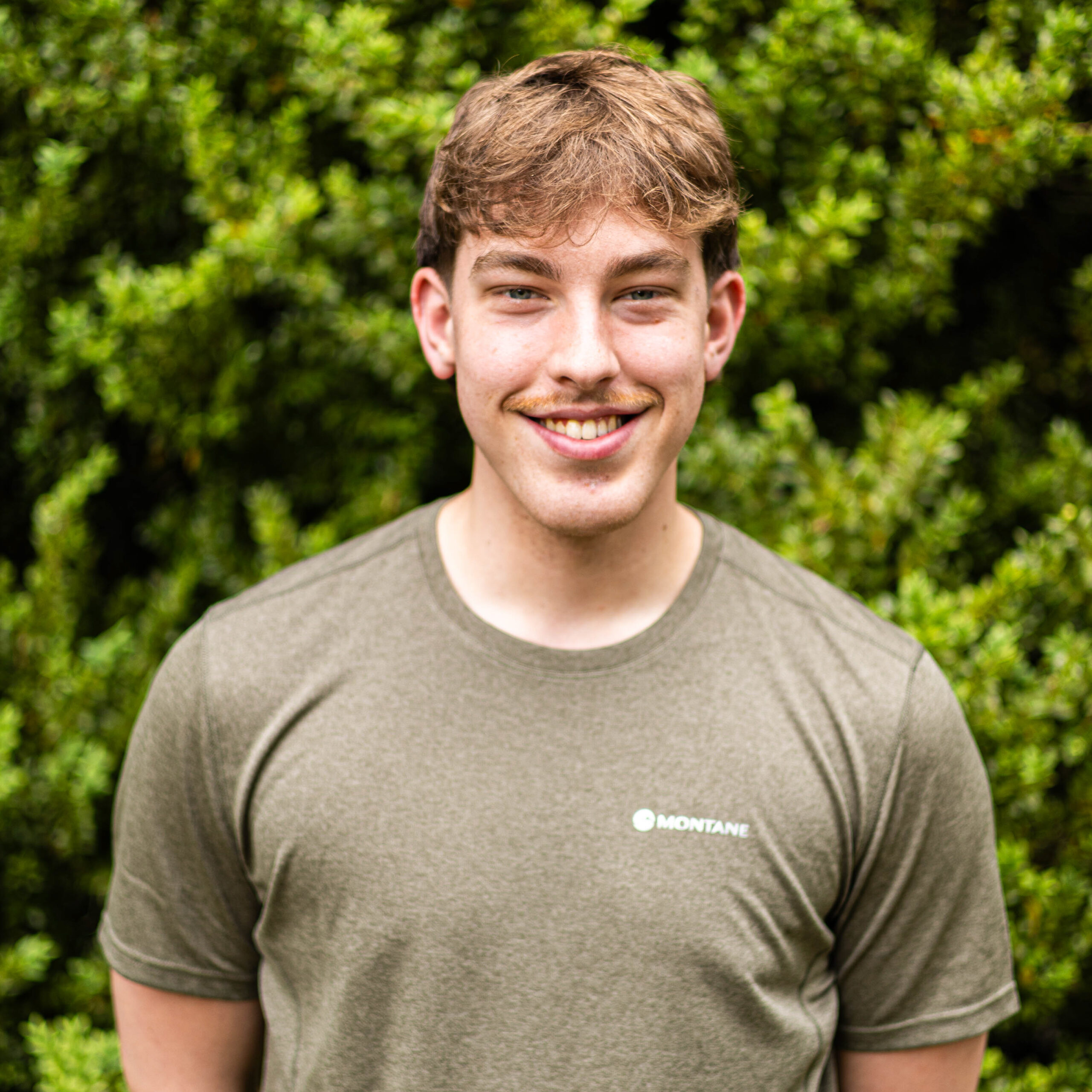 Smiling man wearing an olive-green Montane T-shirt, with green foliage blurred in the background.