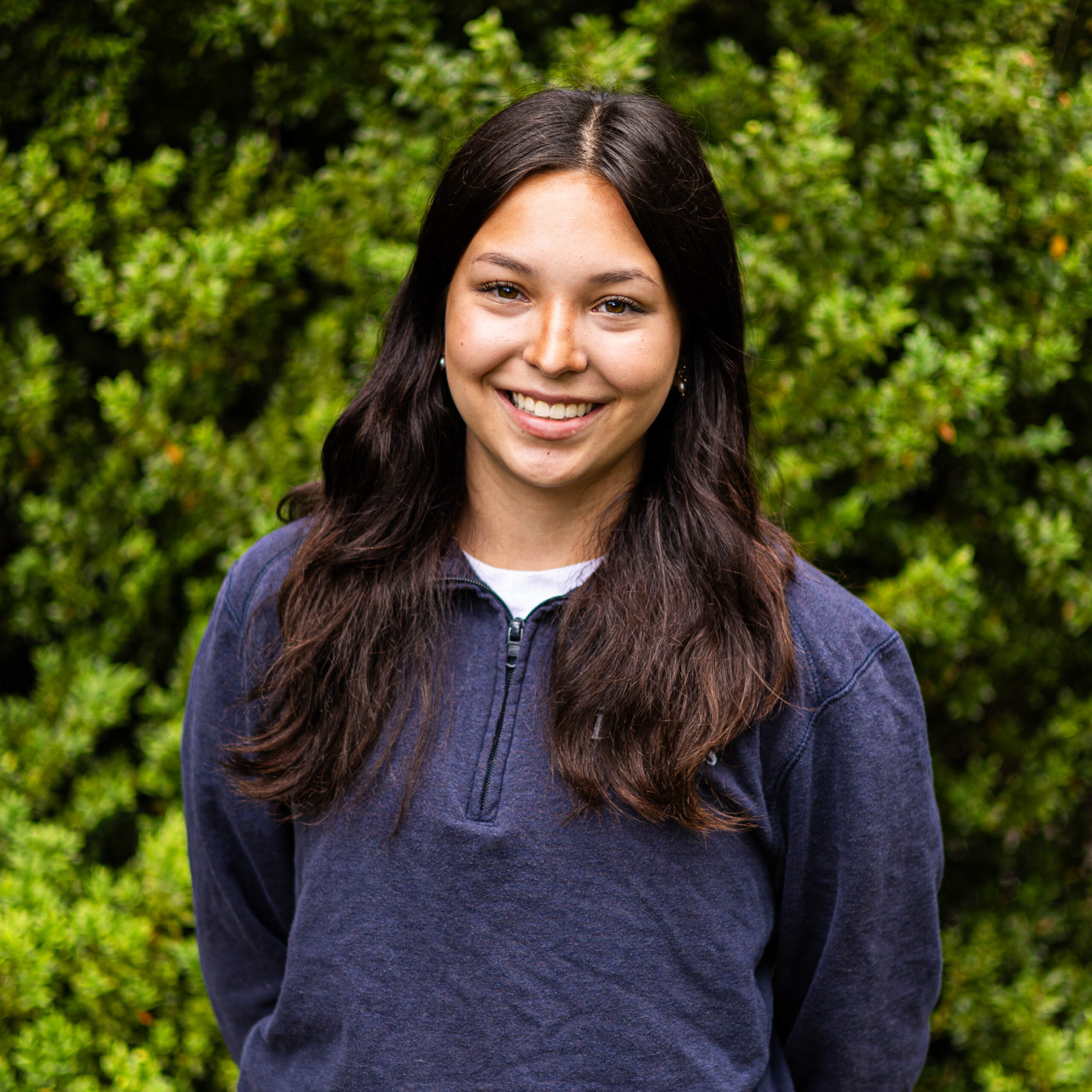 Smiling woman with long dark hair outdoors, green foliage behind her, wearing a dark blue jacket.