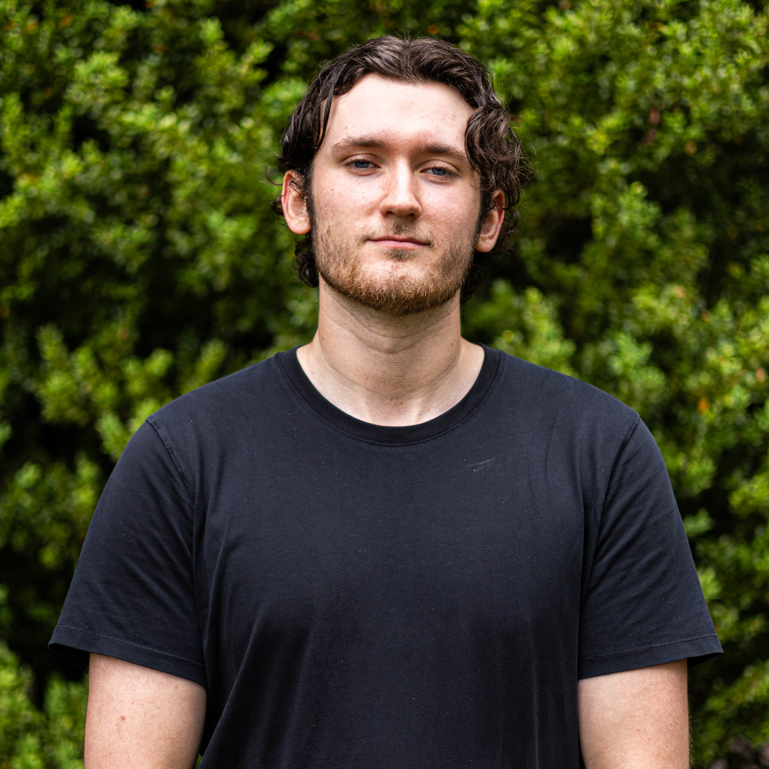 Young man with dark hair and stubble wearing a black t-shirt outdoors, green foliage blurred in the background.