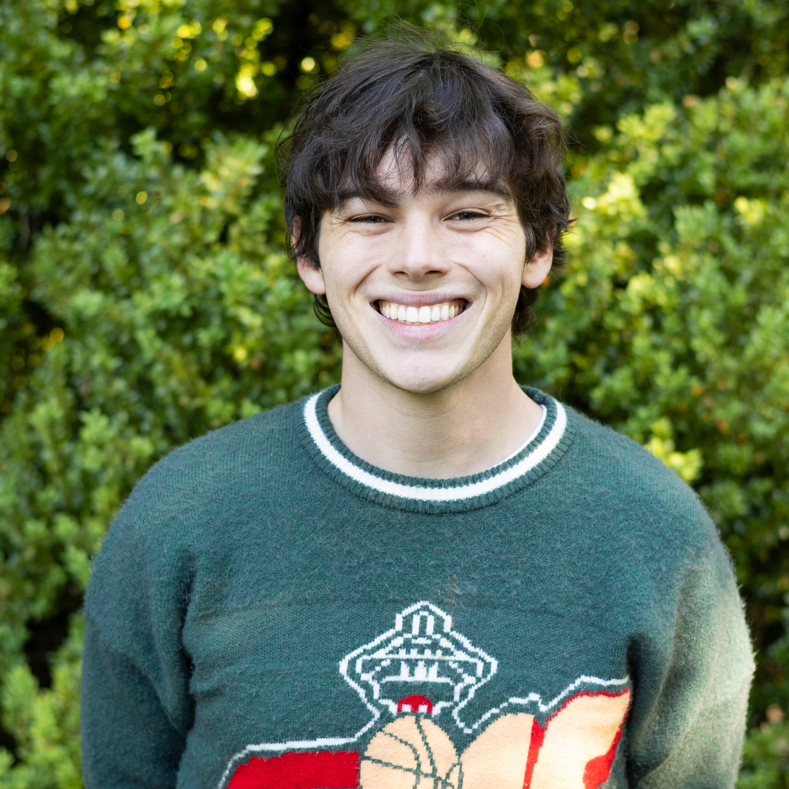 Young man smiling outdoors in a green knitted sweater with a holiday graphic, greenery in the background.