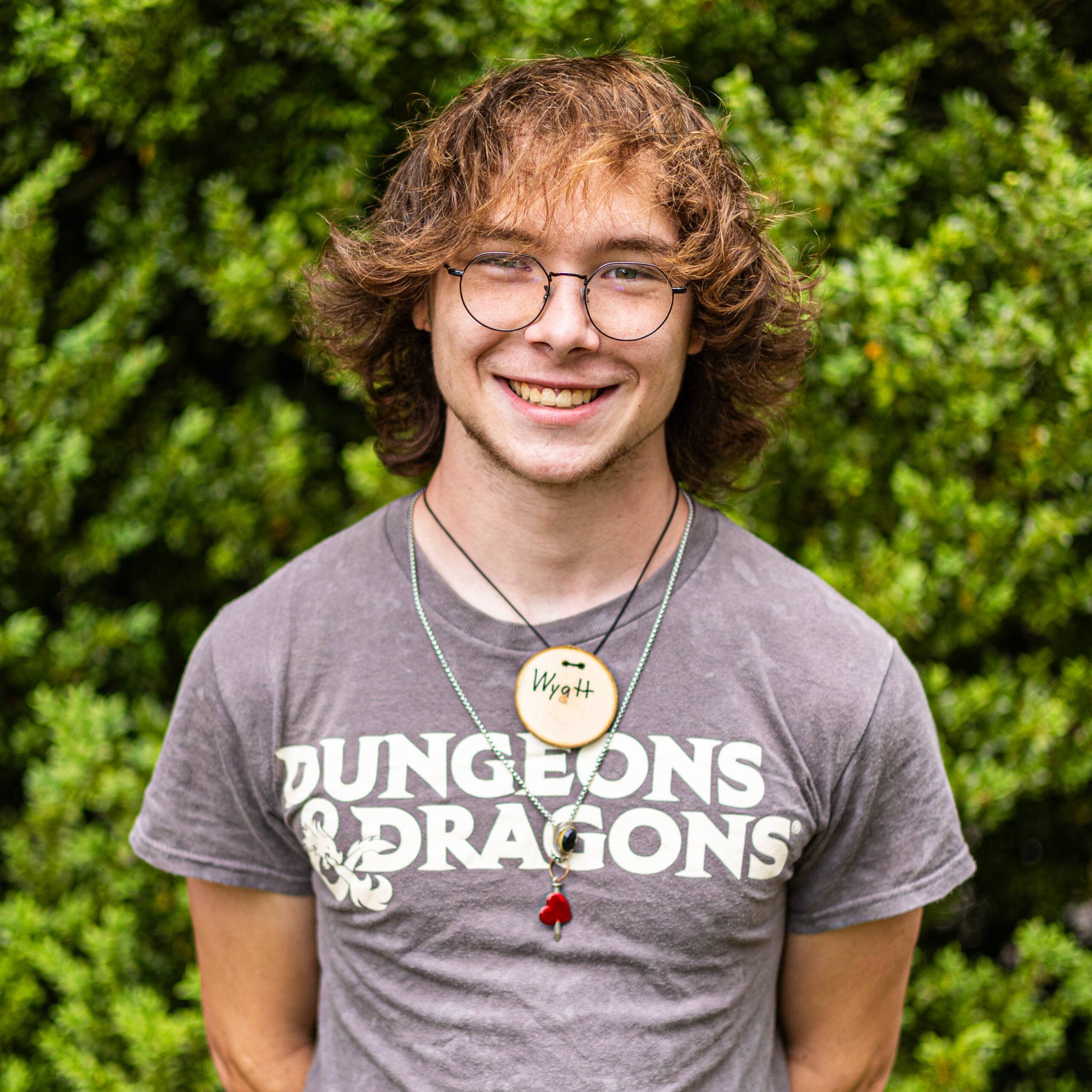 Young person with round glasses and brown hair smiling outdoors, wearing a gray 'DUNGEONS & DRAGONS' T-shirt and necklaces in front of greenery.