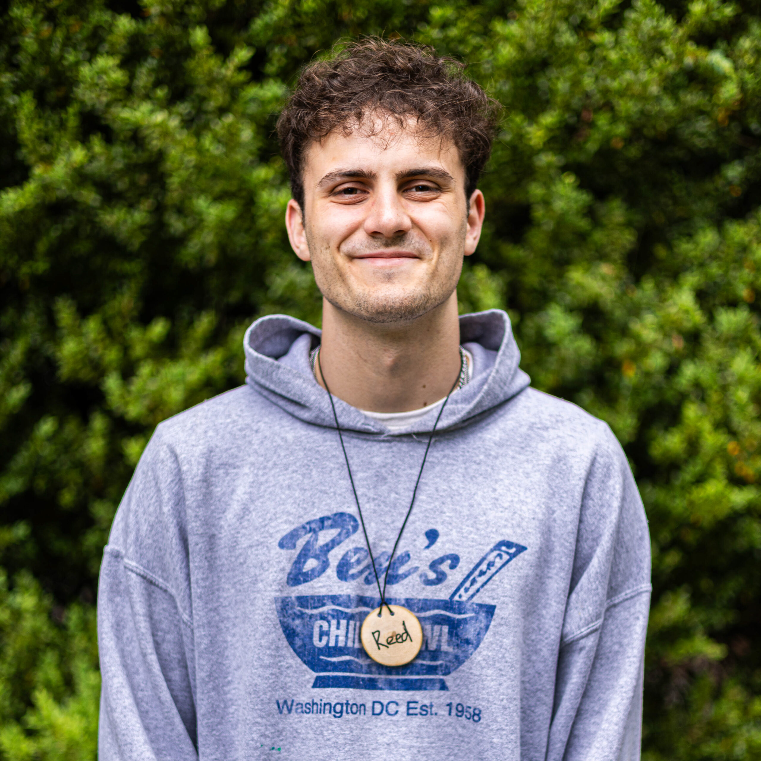 Portrait of a man outdoors, smiling, wearing a gray hoodie with a round wooden tag reading 'Reed' on a cord, green bushes in the background.