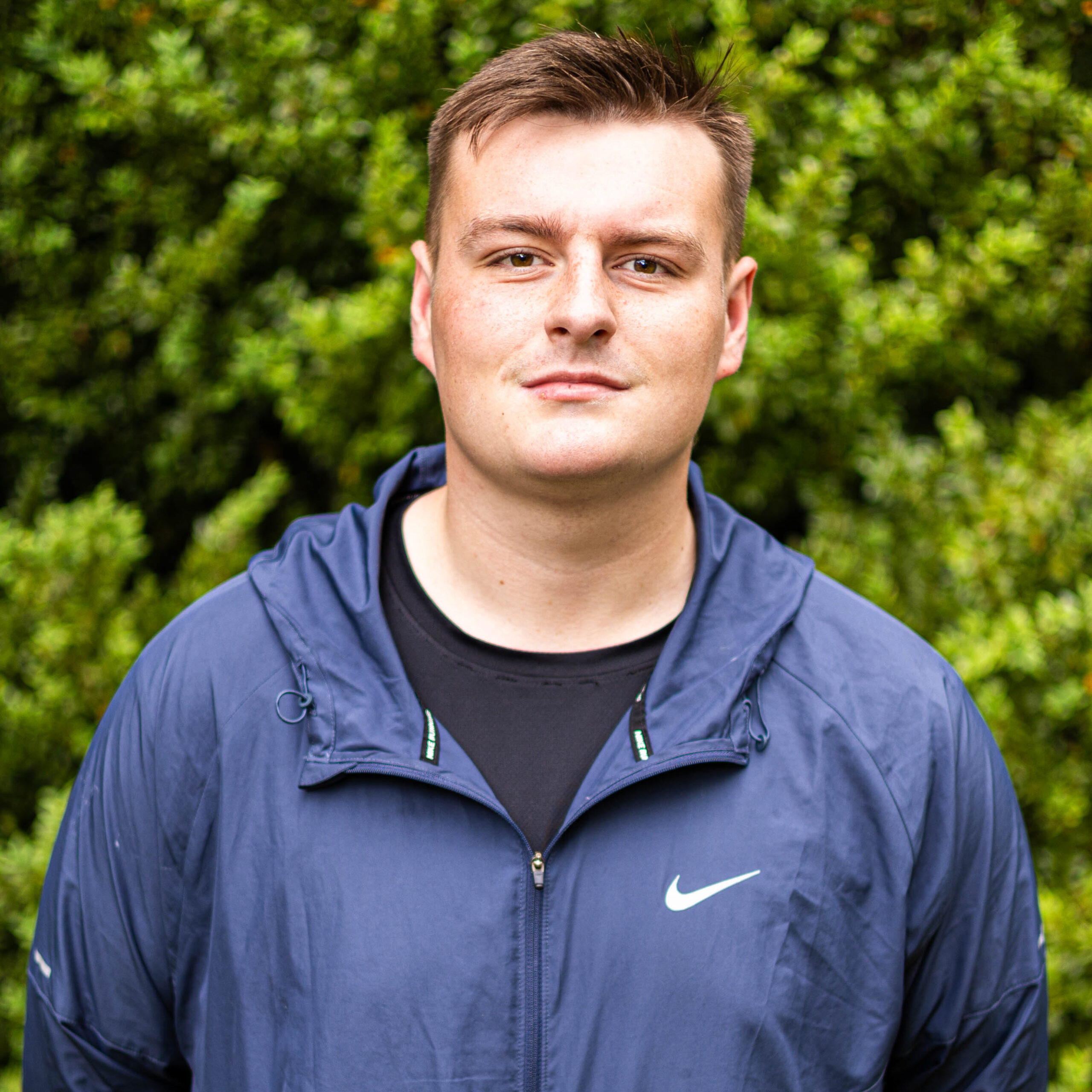 Portrait of a man in a blue jacket outdoors with green foliage behind him.