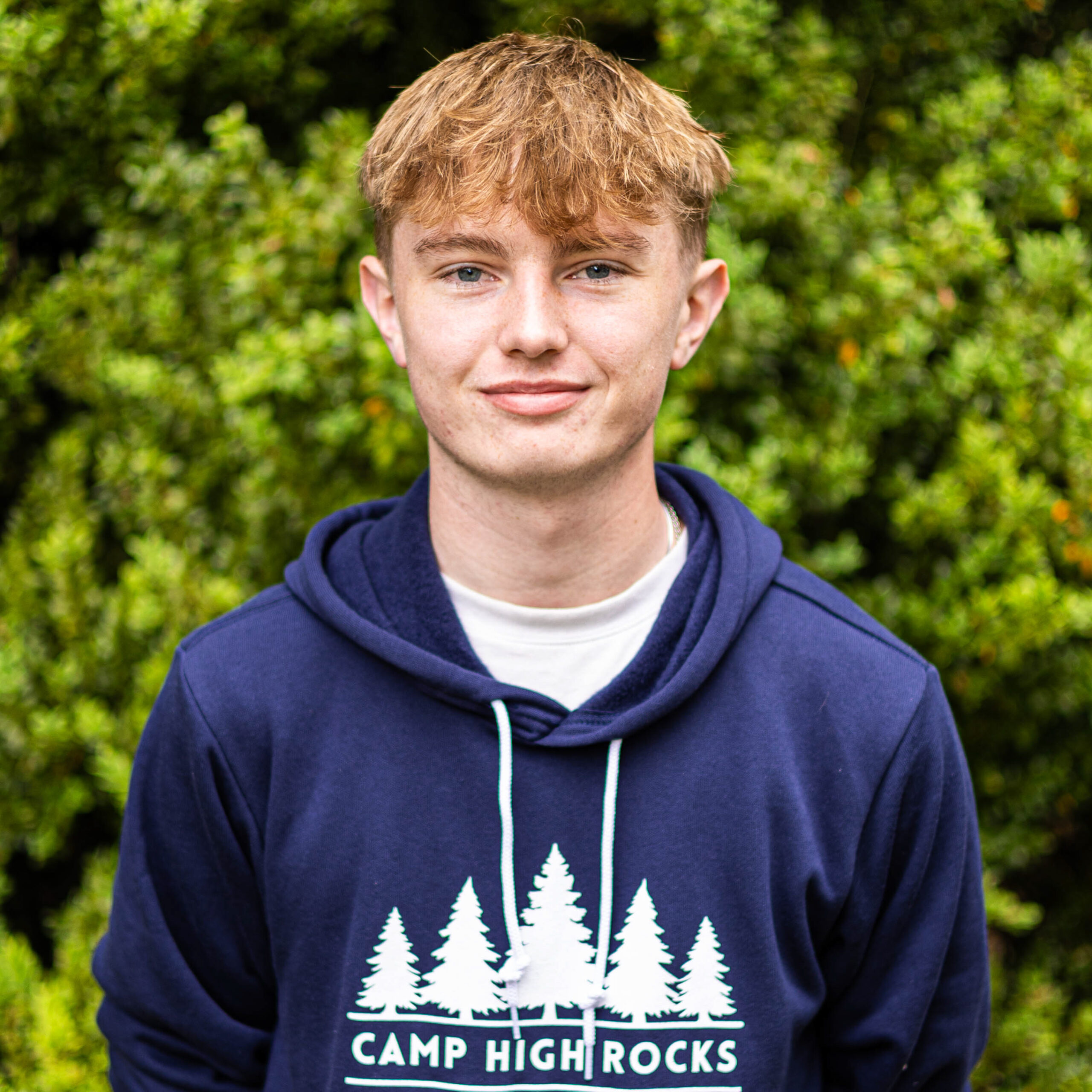 Young man wearing a navy hoodie with white pine-tree graphics and 'CAMP HIGH ROCKS' text, standing outdoors with green foliage in the background.