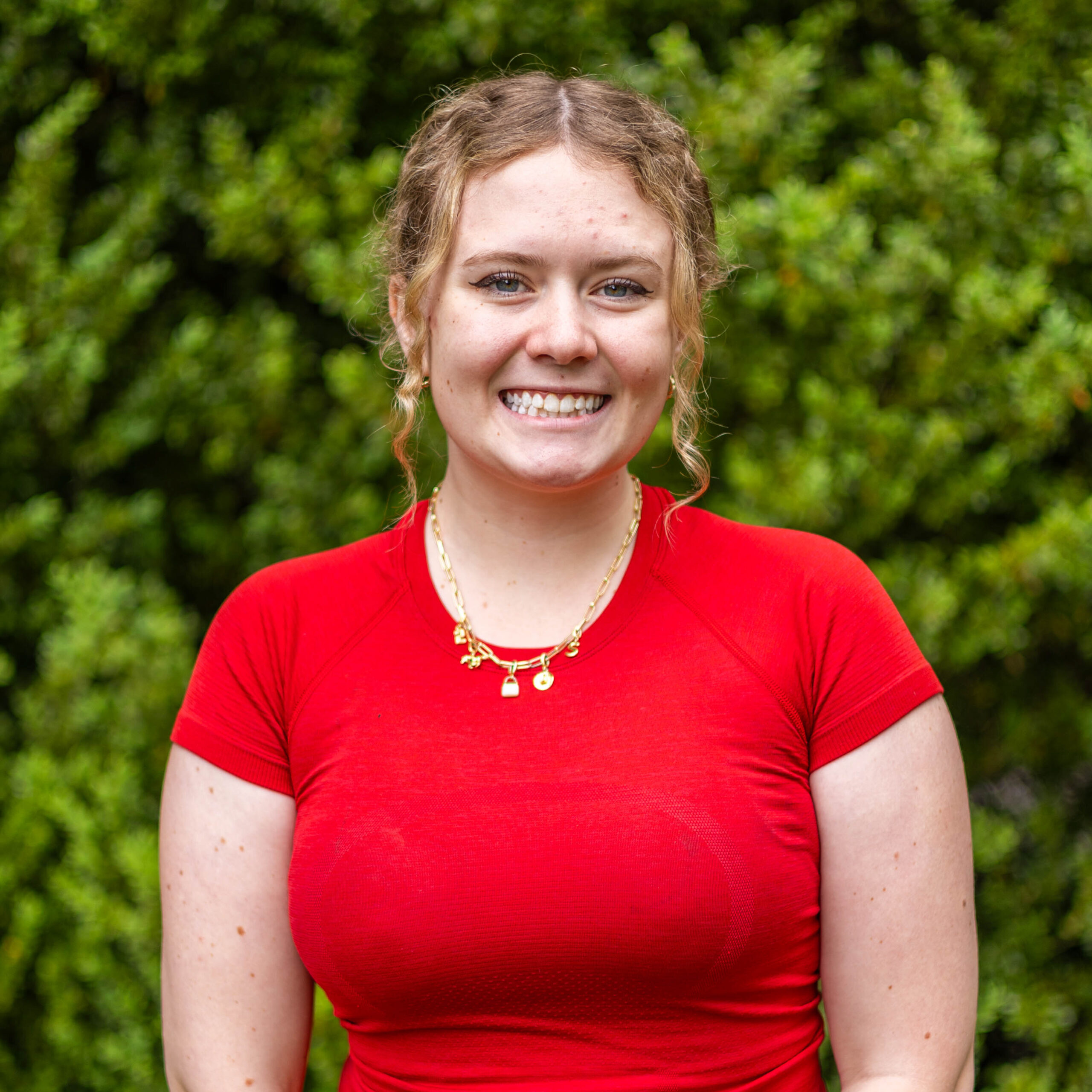 Smiling woman in a red shirt outdoors with a green leafy background.
