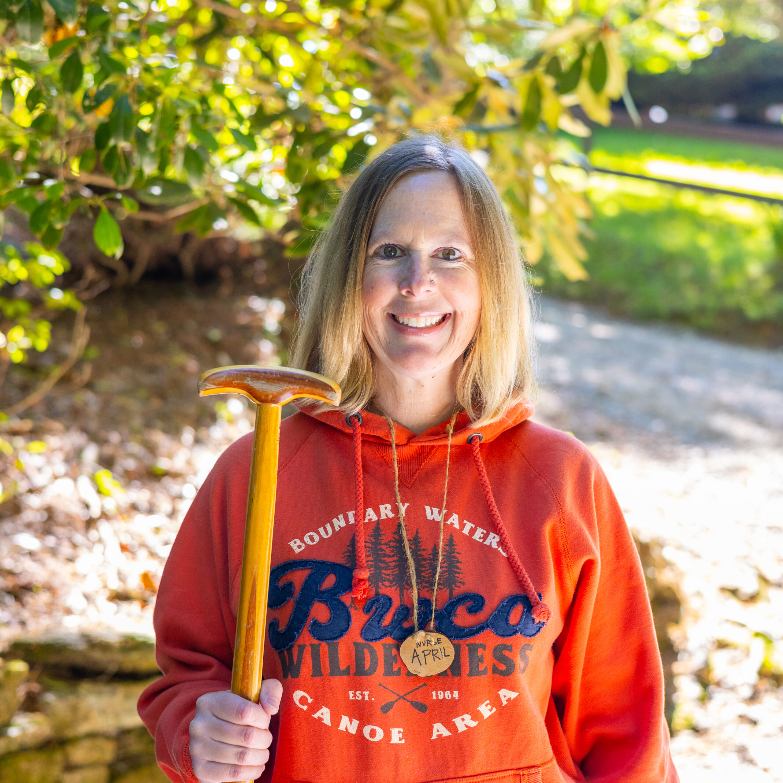 Smiling person outdoors in an orange hoodie holding a wooden hiking staff, with trees and sunlight in the background.