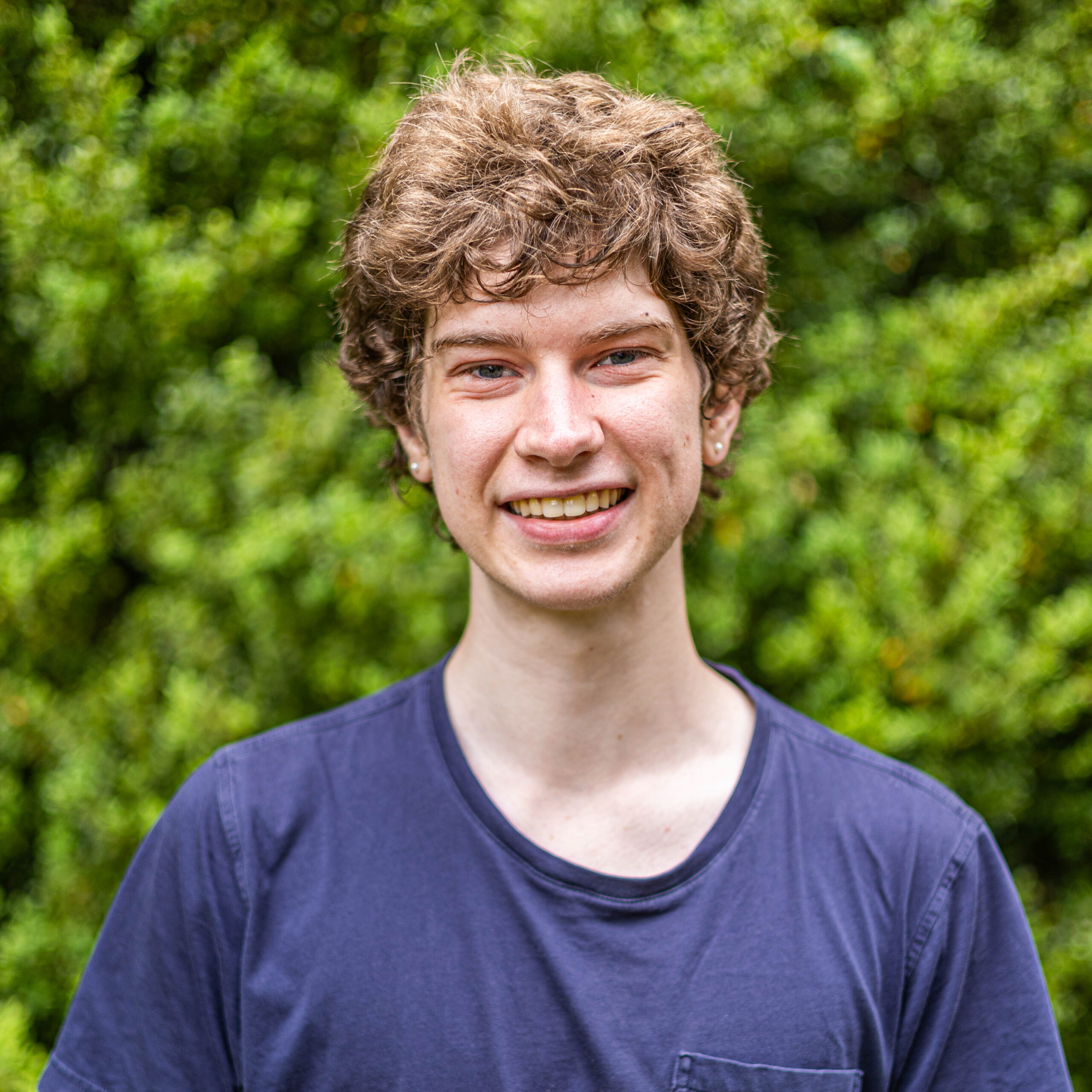 Close-up of a smiling person wearing a dark blue T-shirt outdoors with a green leafy background.