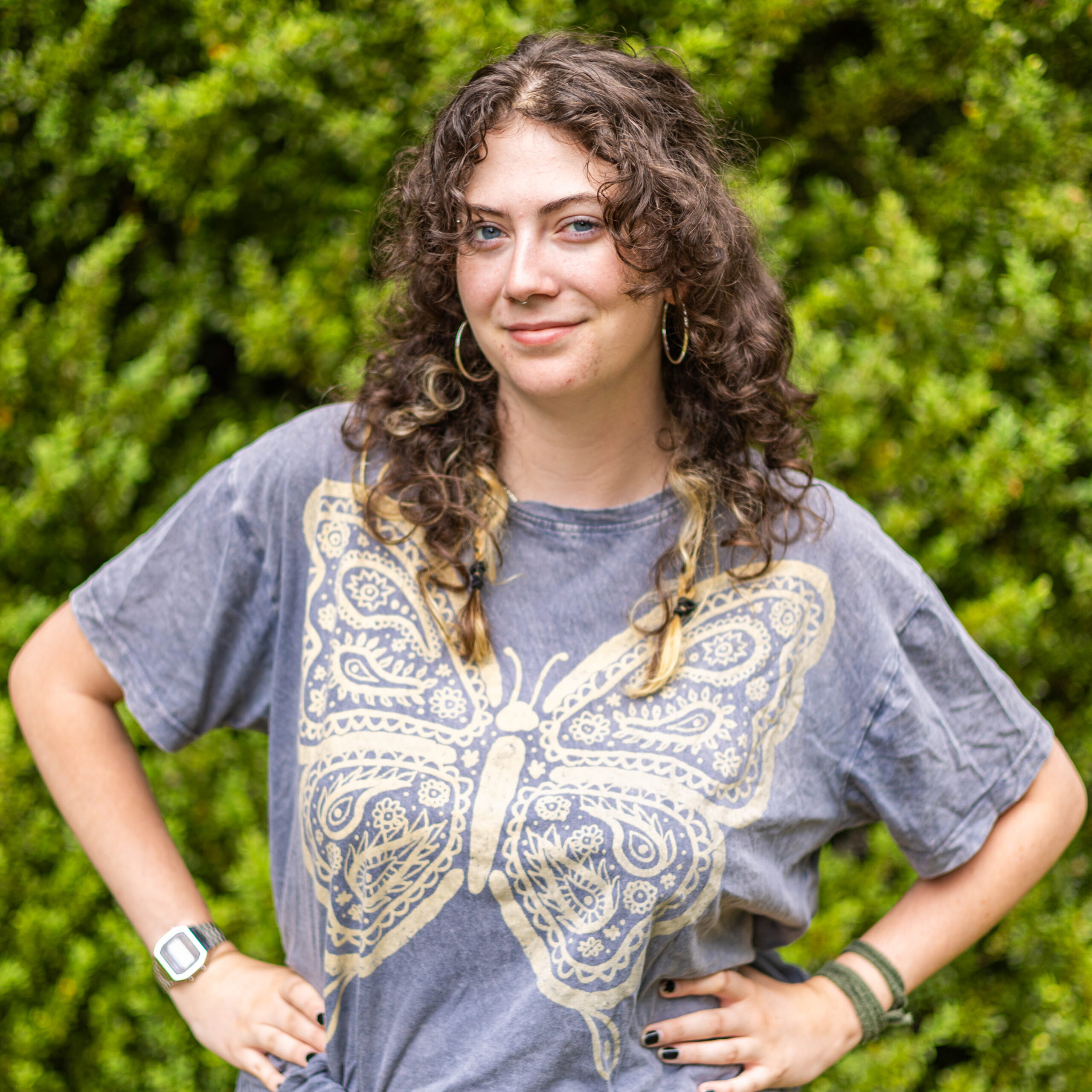 Woman with curly brown hair wearing a blue-gray patterned shirt, standing outdoors with green bushes in the background, hands on hips and a slight smile.