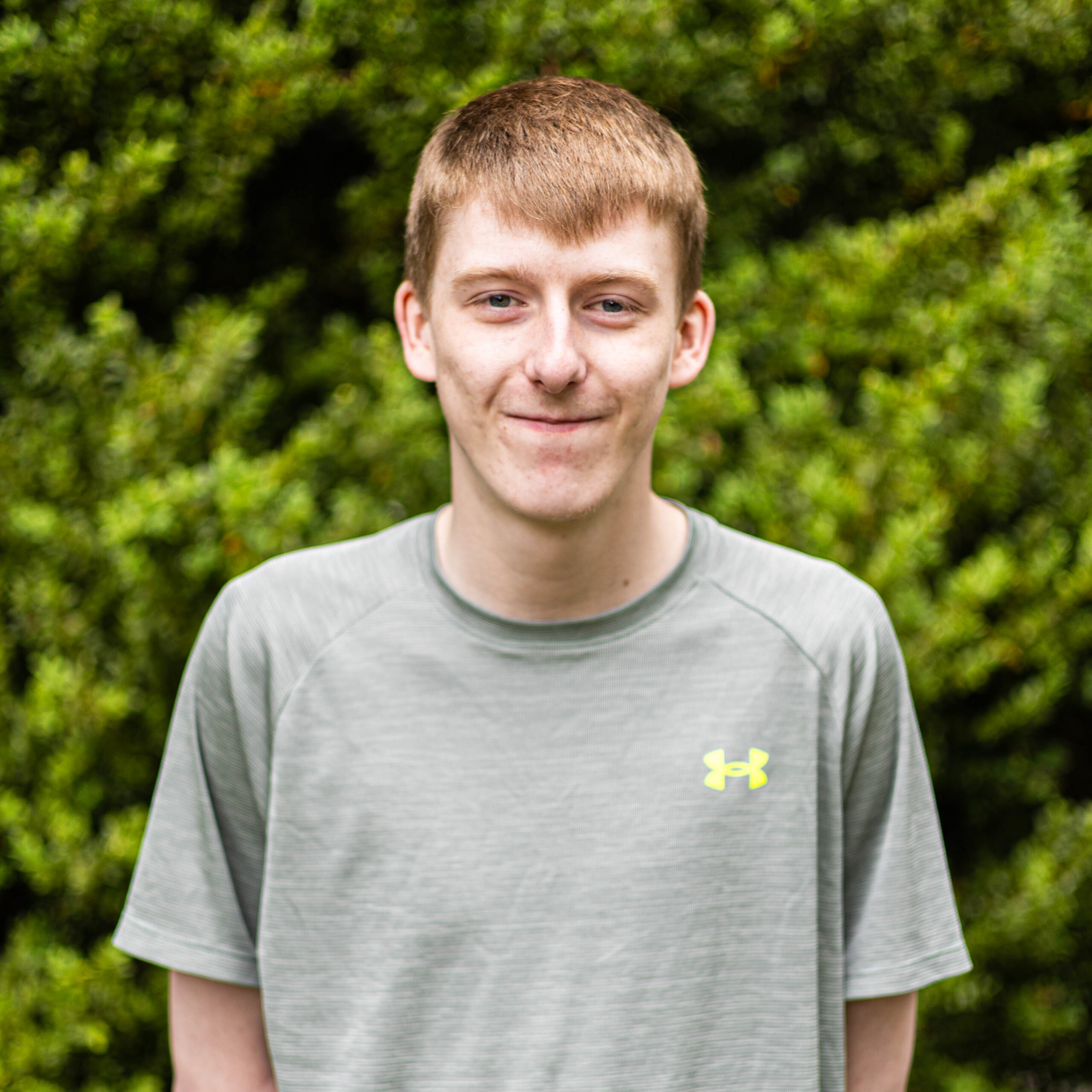 Young man in a gray athletic shirt outdoors with green bushes in the background, facing the camera.