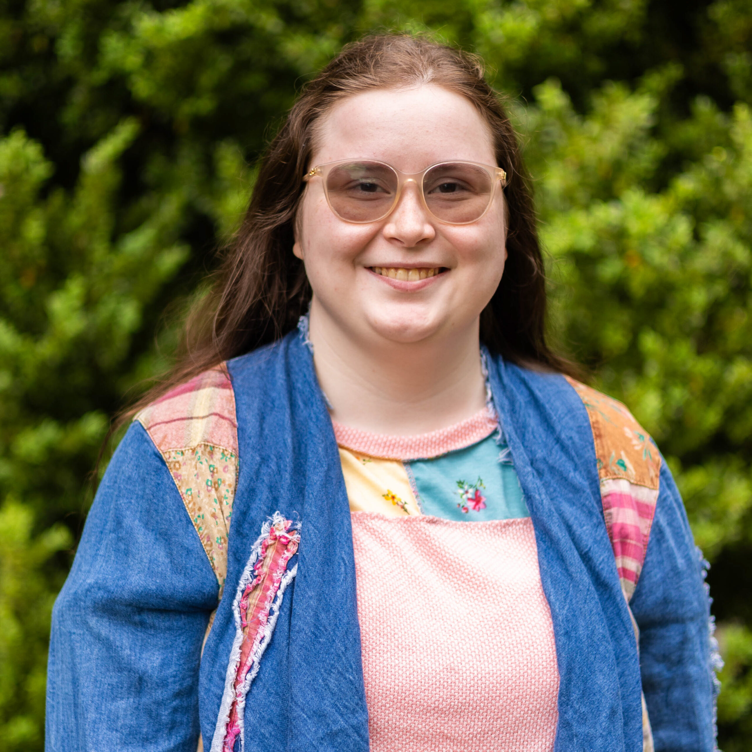 Close-up of a smiling woman wearing large translucent pink sunglasses and a patchwork denim jacket, with green foliage behind her.