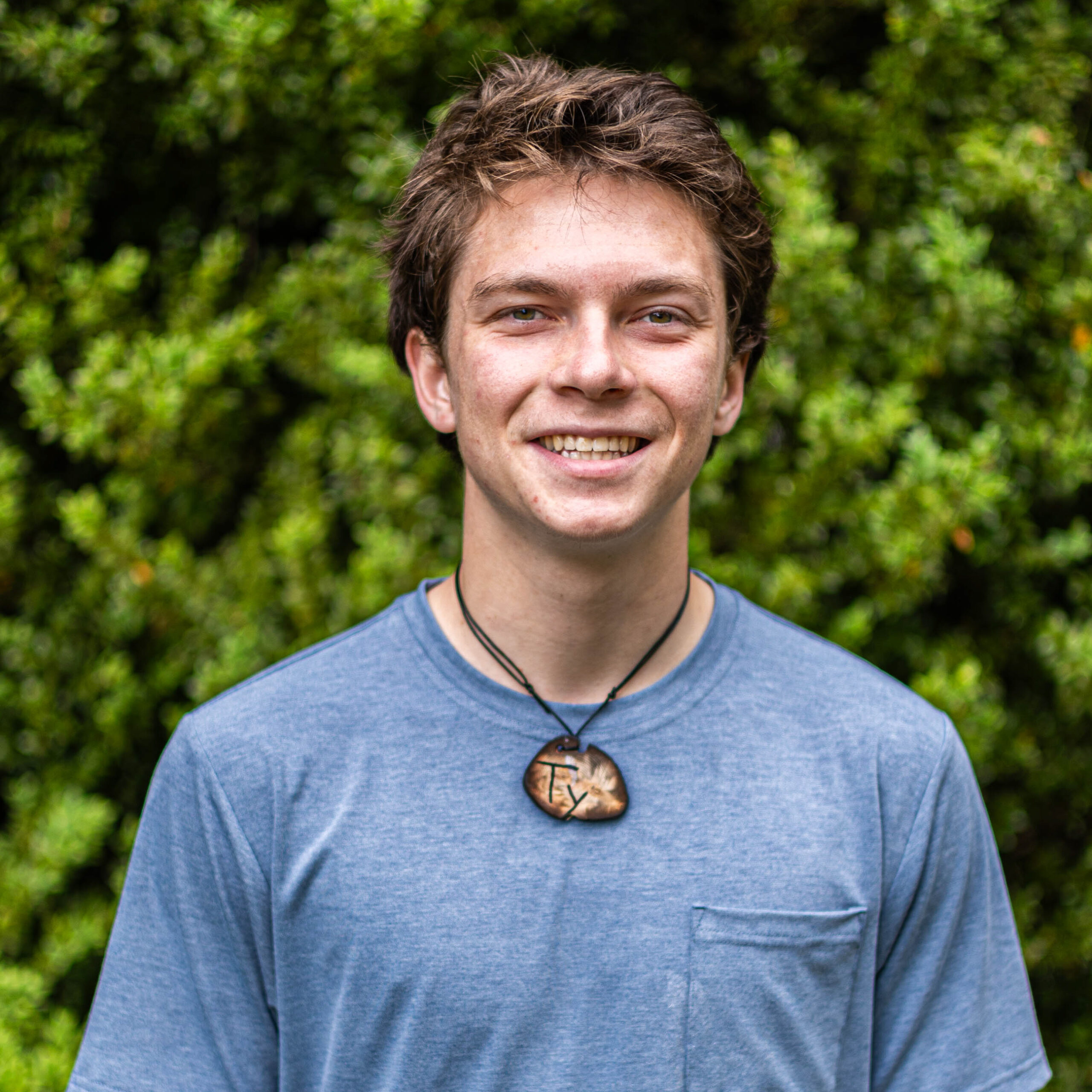 Close-up of a smiling man wearing a blue shirt and a black necklace, with green foliage in the background outdoors.