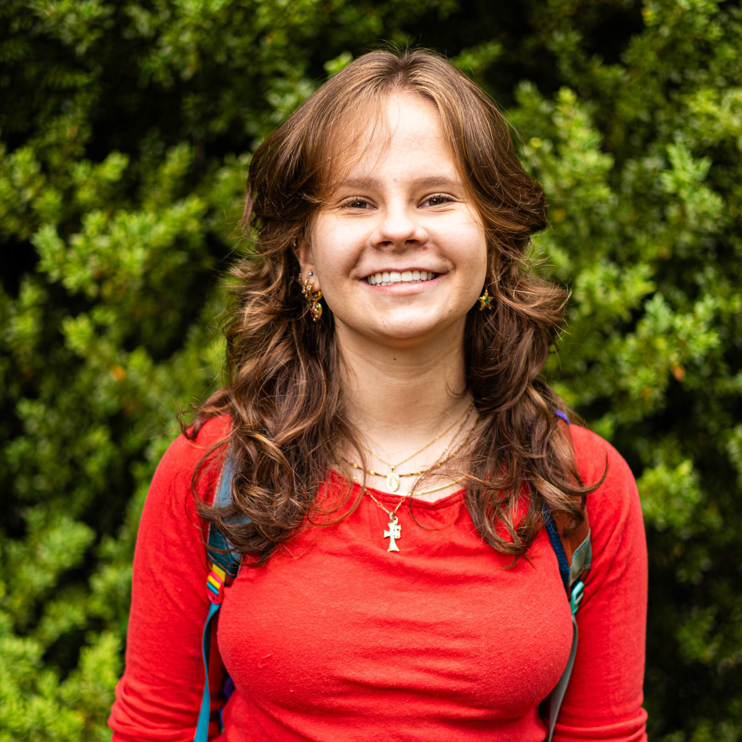 Smiling woman outdoors in a red sweater with a gold cross necklace, green bushes in the background.