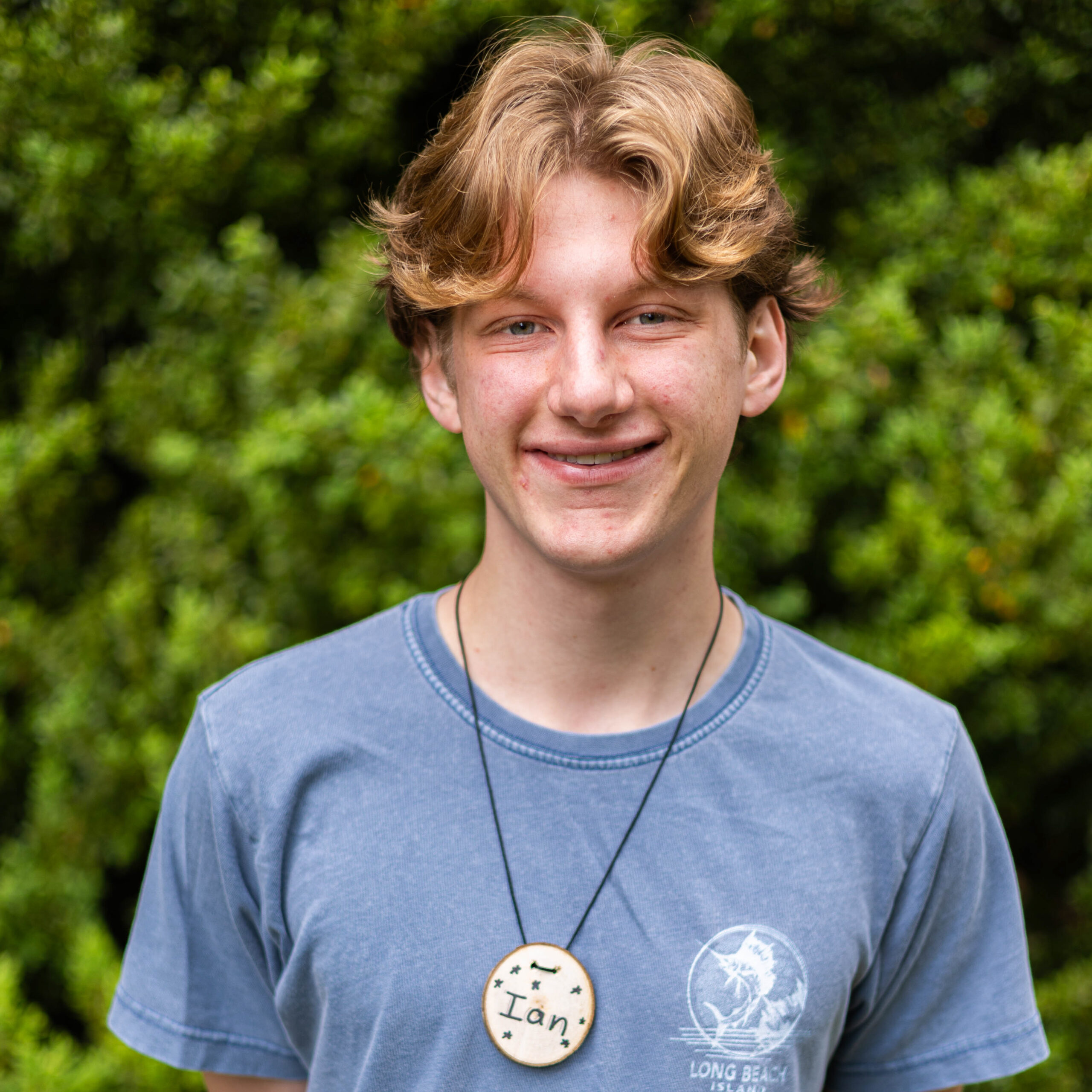 Smiling young man outdoors in a blue shirt with two circular badges, one reading 'Ian'; green bushes in the background.