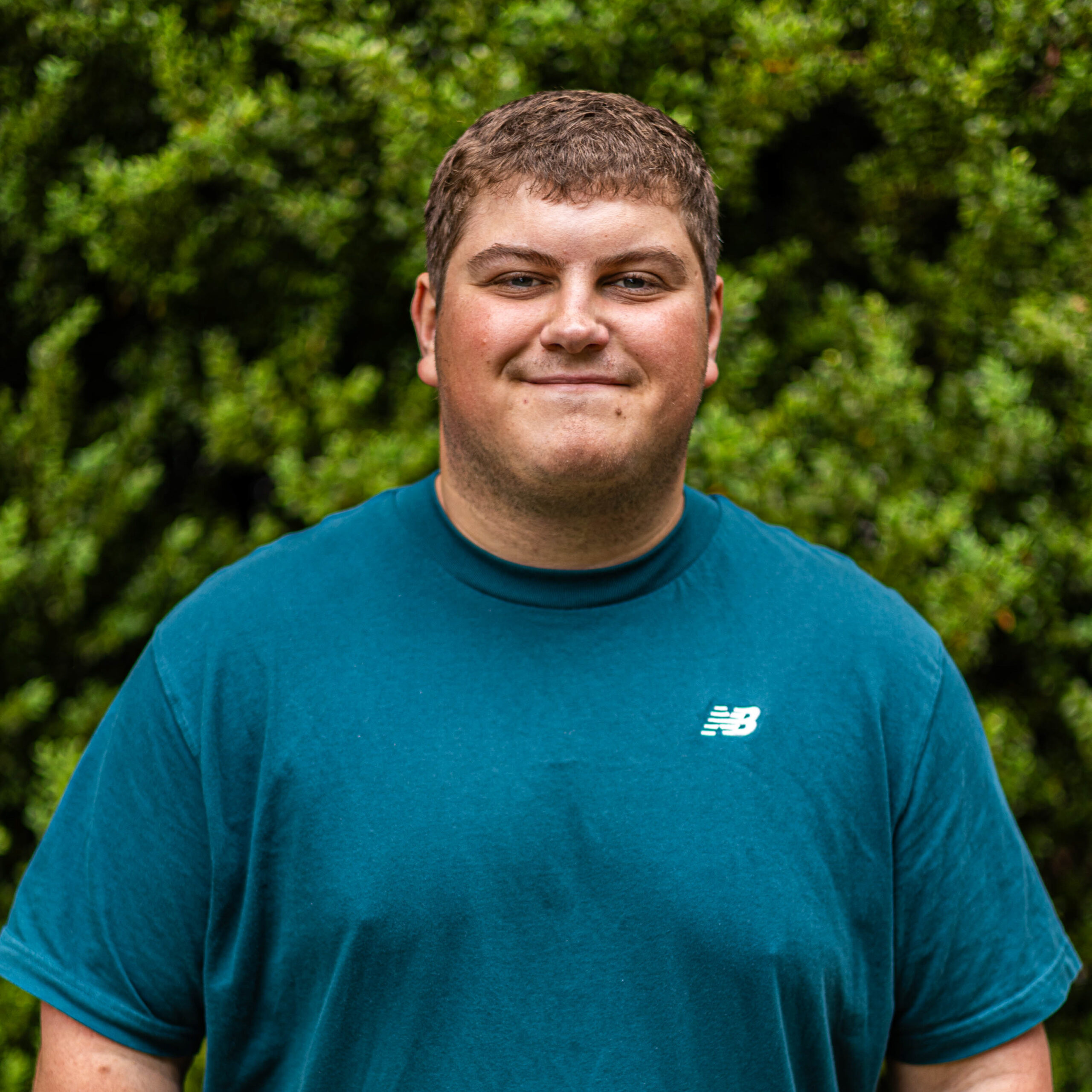 Man wearing a teal T-shirt outdoors with dense green foliage in the background.