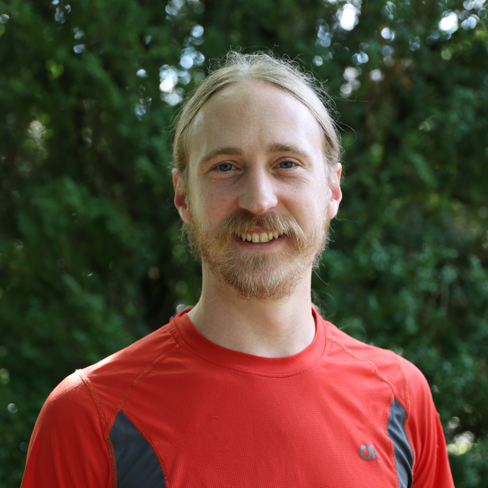 Smiling man with blond hair and a beard outdoors in a red athletic shirt.