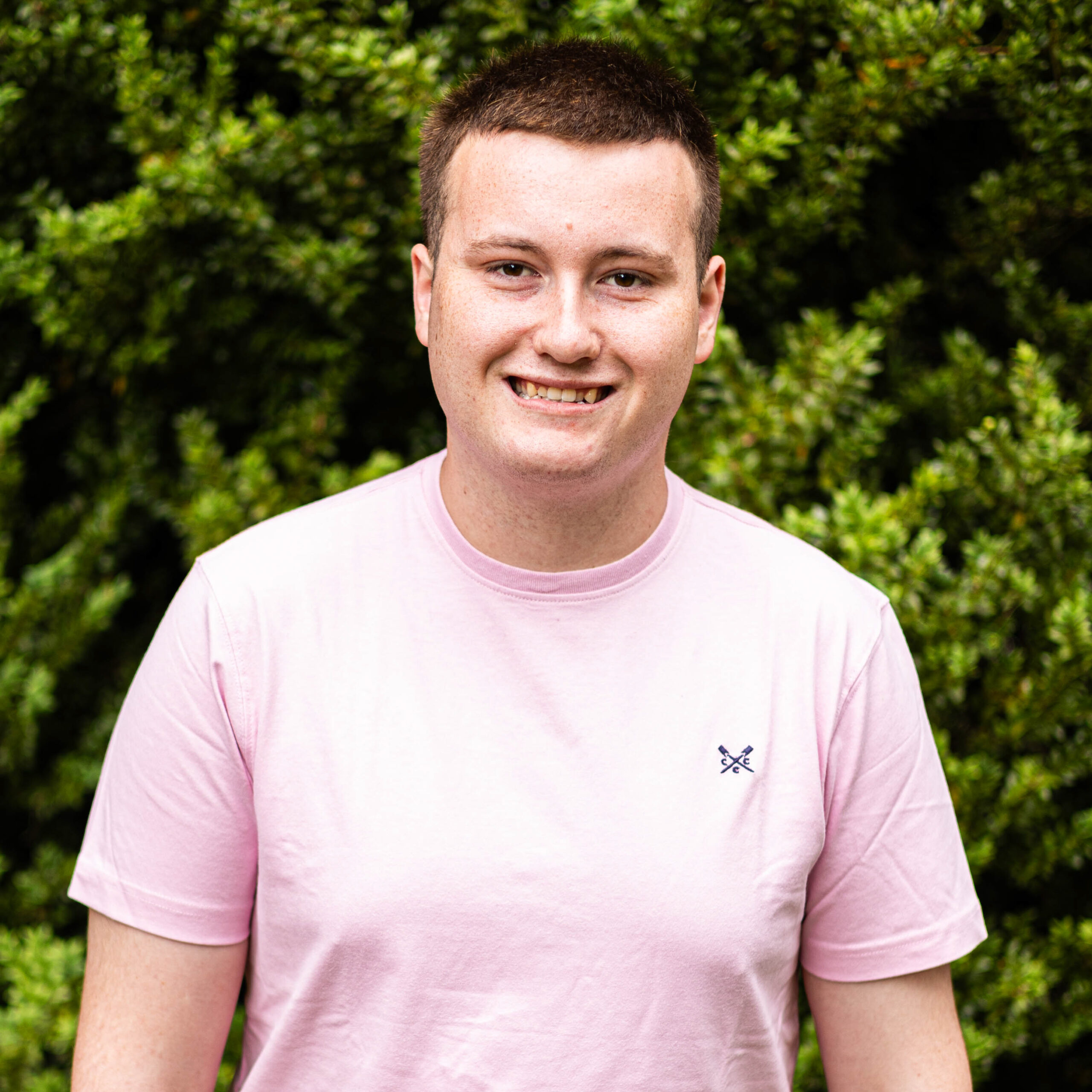 Smiling man in a light pink T-shirt outdoors with green bushes in the background.