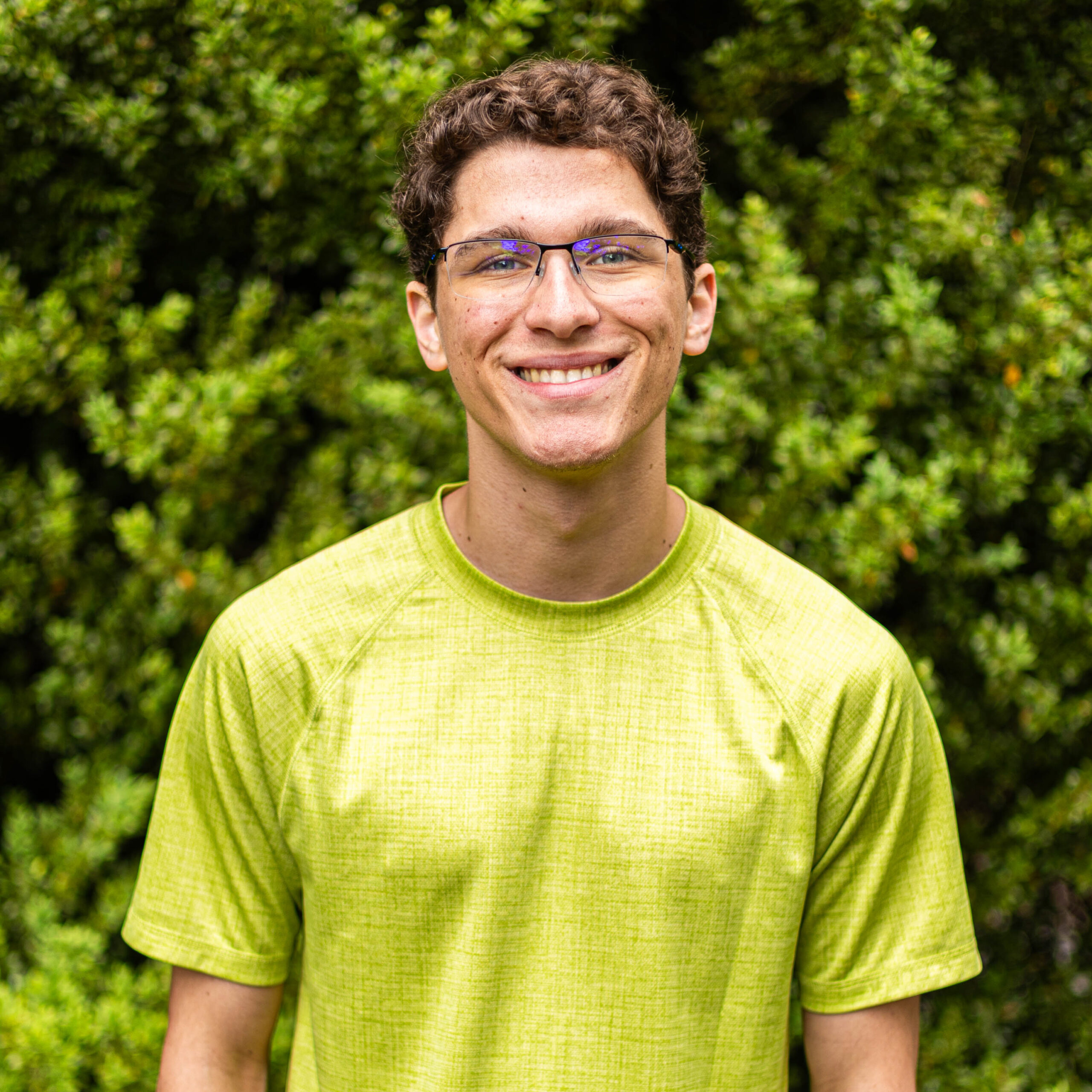 Smiling young man wearing glasses and a bright lime shirt outdoors with greenery in the background.
