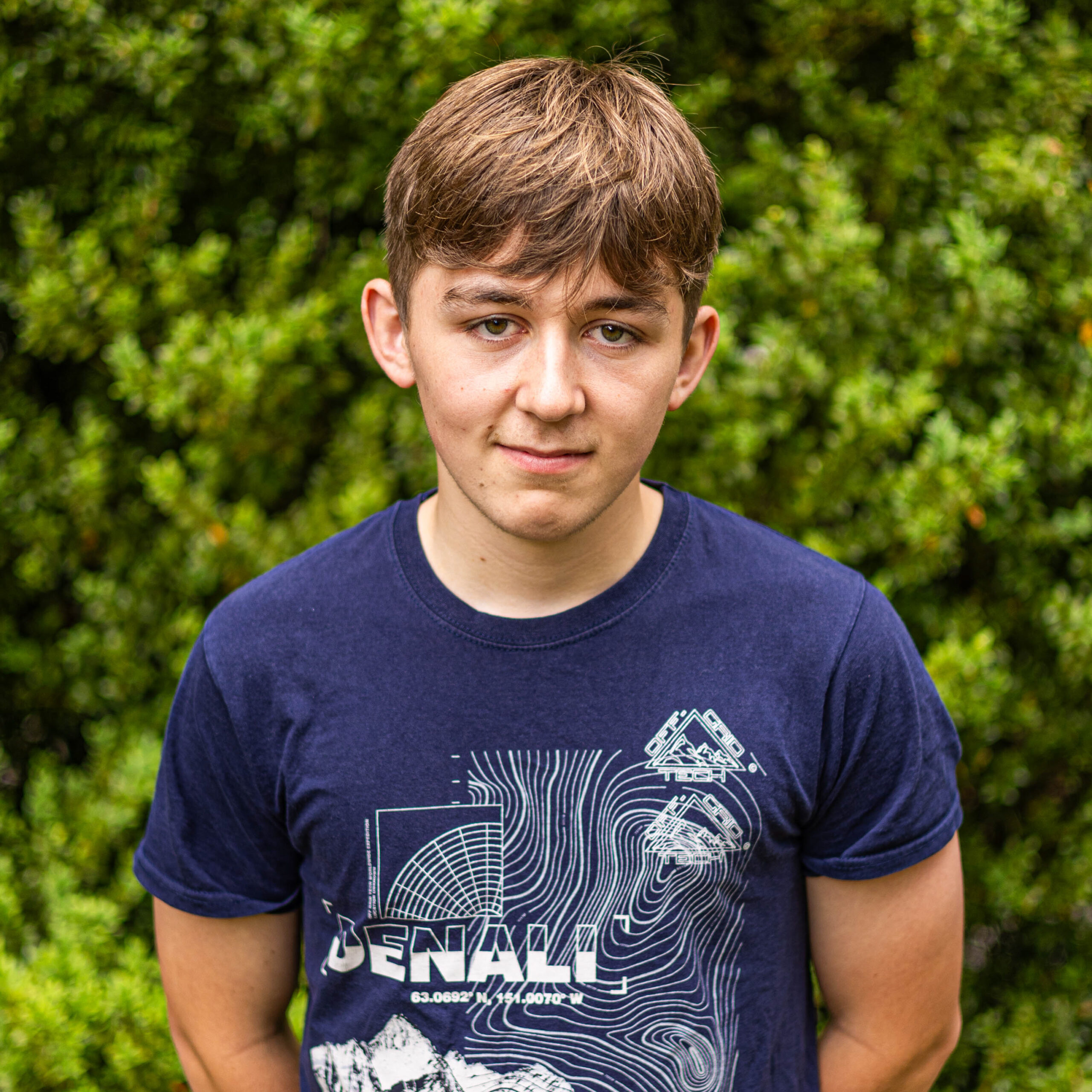 Young man wearing a navy T-shirt with white geometric graphics, standing outdoors with green bushes in the background.