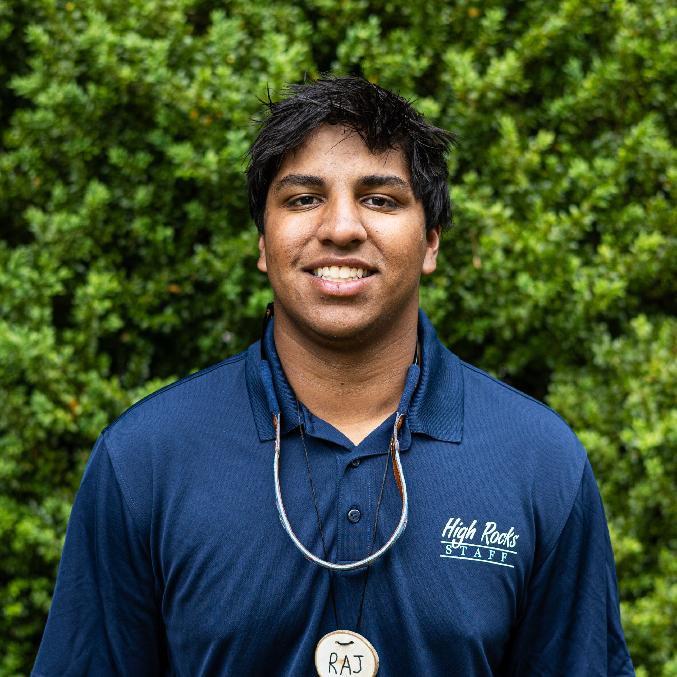 Outdoor portrait of a smiling man in a navy shirt, sunglasses hanging from his neck, with a green leafy background and a 'High Rocks STAFF' logo on the chest.