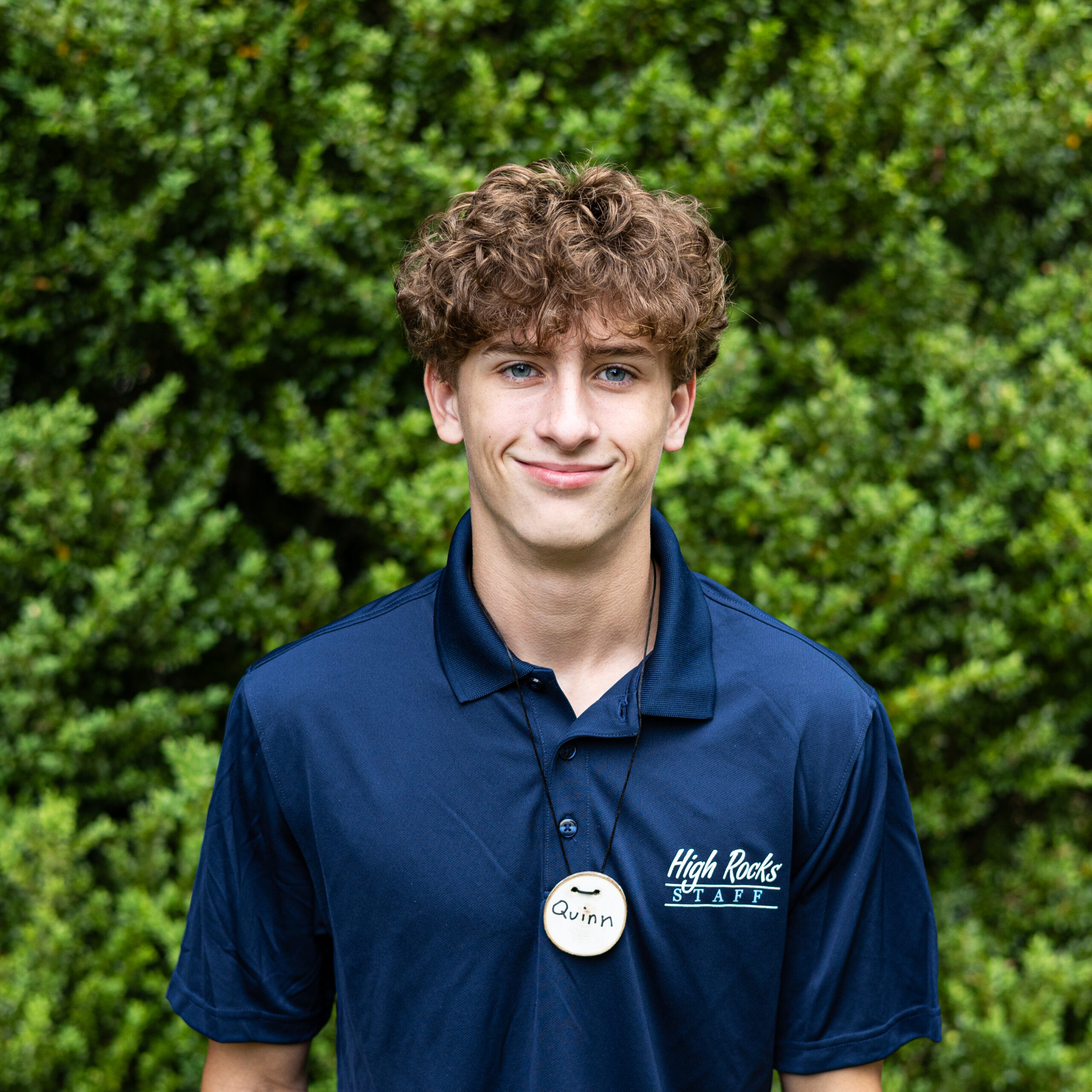 Young man with curly brown hair in a navy High Rocks staff polo, name badge Quinn, outdoors with a green hedge background.