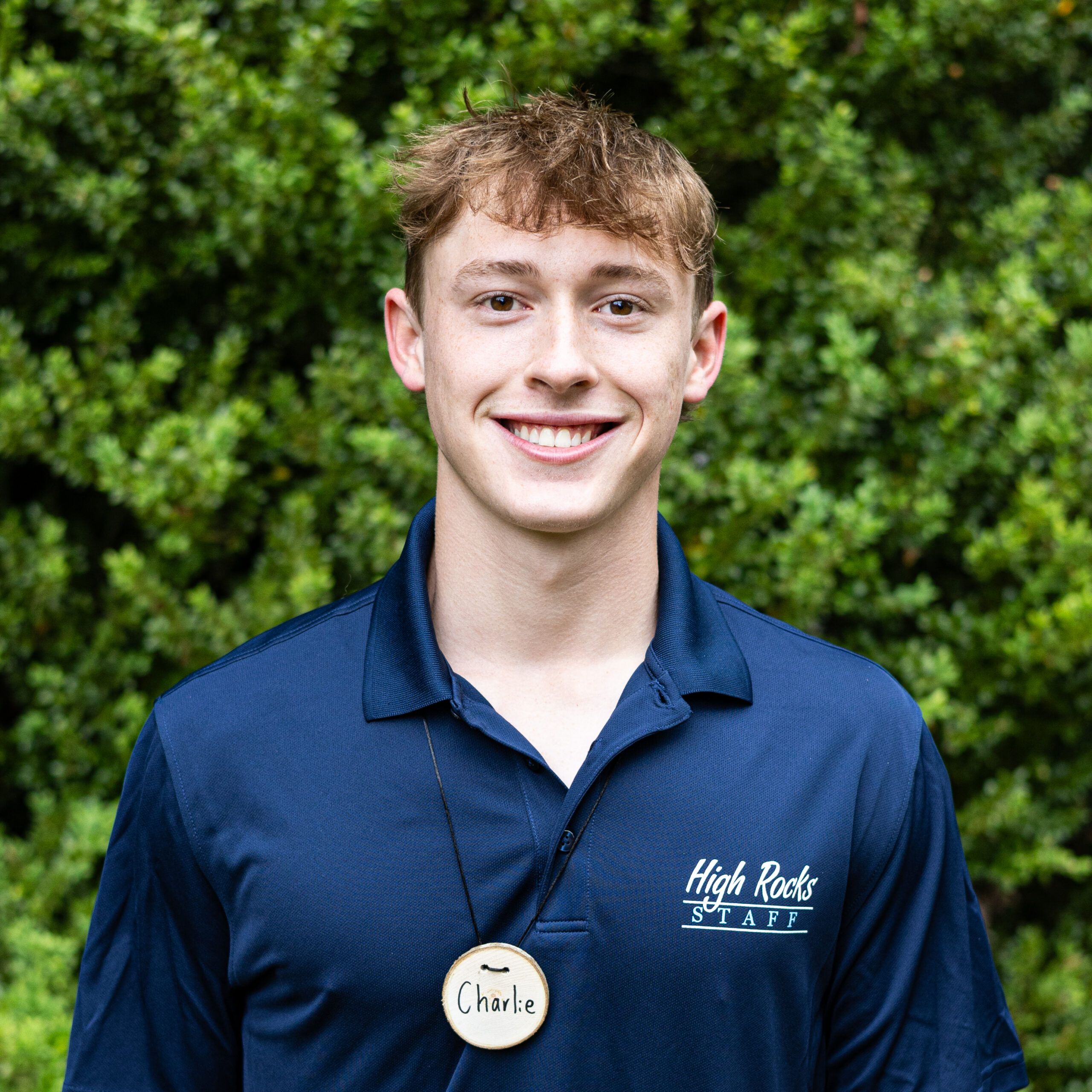 Portrait of a young man outdoors in a navy polo with a High Rocks STAFF logo and a round wooden name badge reading ‘Charlie’ hanging at his chest