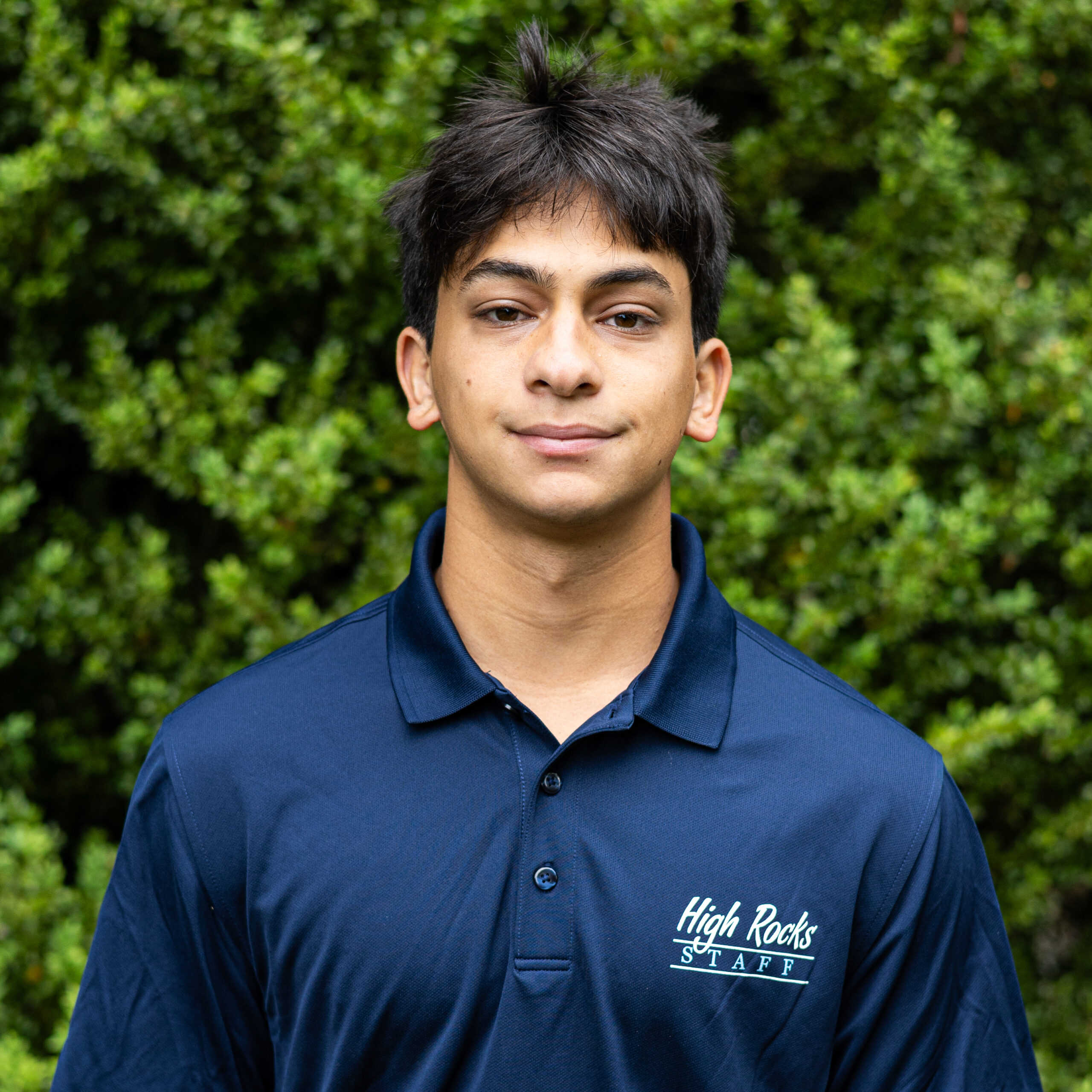 Close-up portrait of a young man wearing a navy polo with a 'High Rocks STAFF' logo, outdoors with green foliage in the background.
