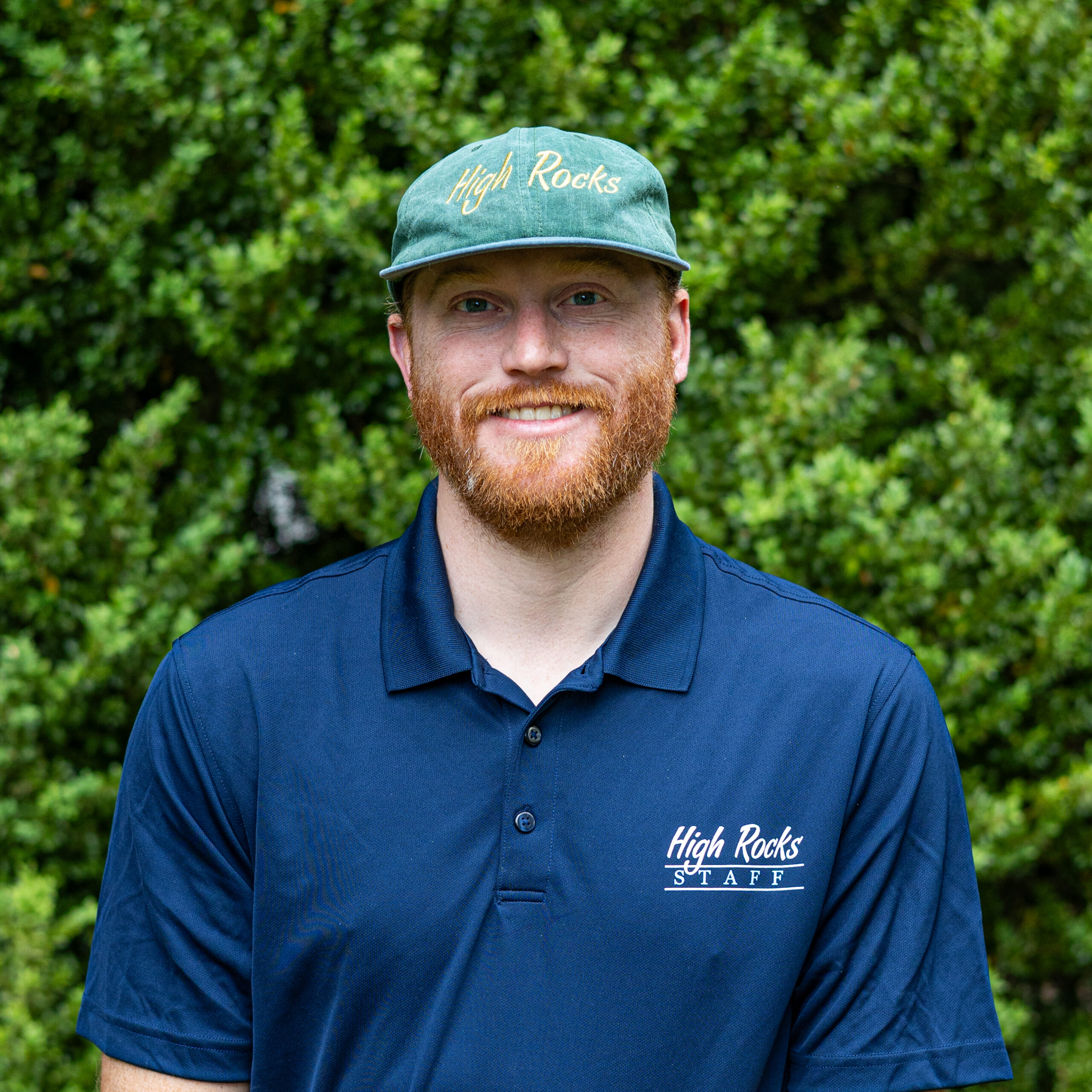 Smiling man wearing a navy polo with the 'High Rocks STAFF' logo, outdoors with green foliage in the background.