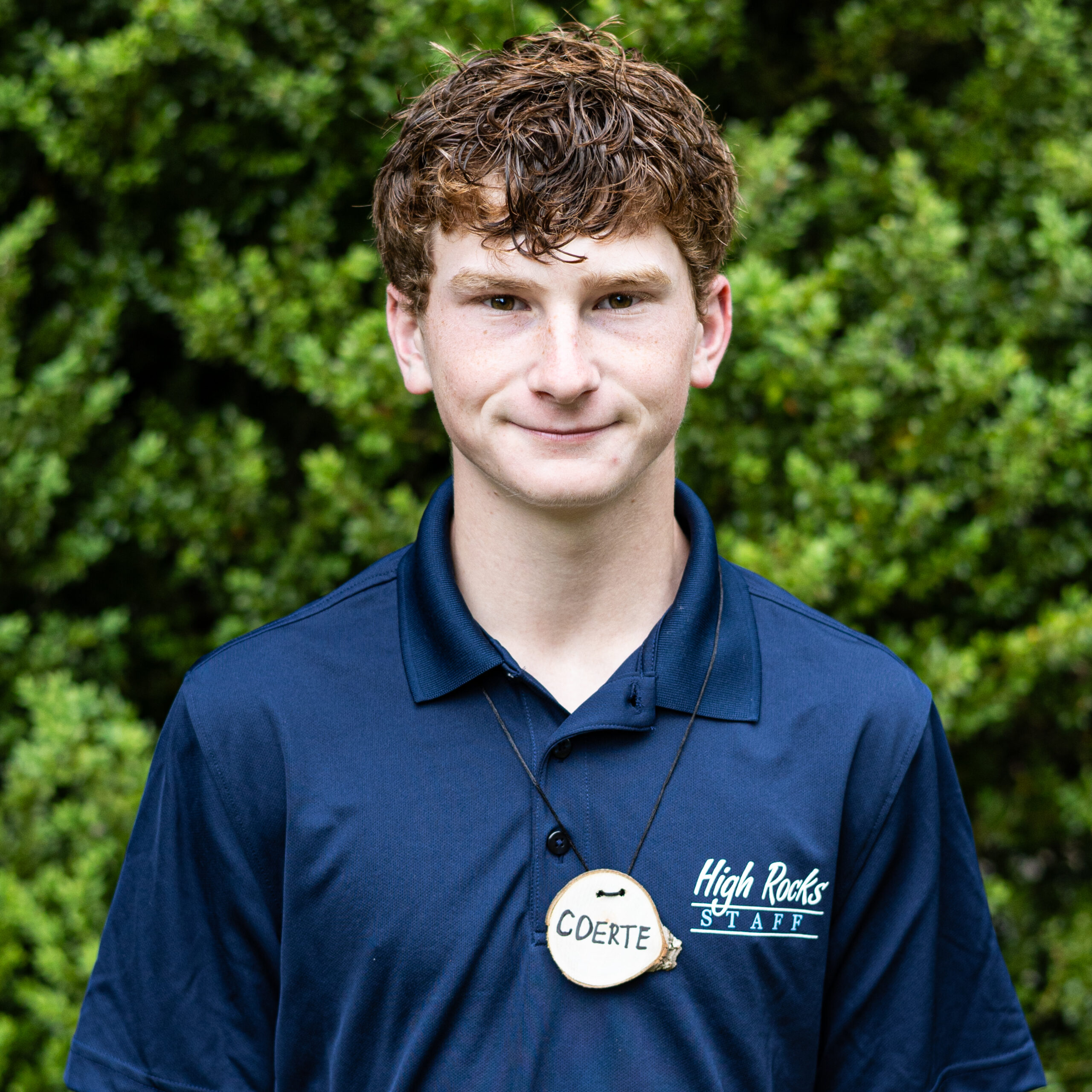 Young man in a navy polo with a round name badge reading 'COERTE' and 'High Rocks STAFF' on chest, outdoors with green bushes in background.