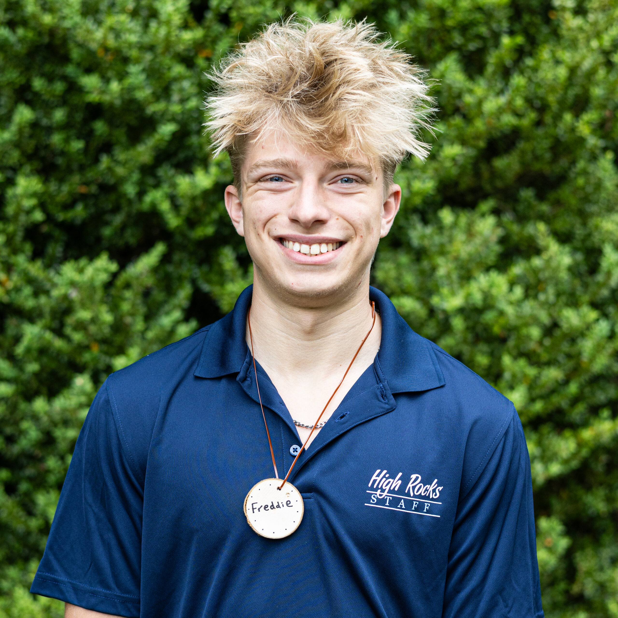 Smiling young man with messy blond hair outdoors, wearing a blue High Rocks staff polo and a round name badge that reads Freddie.