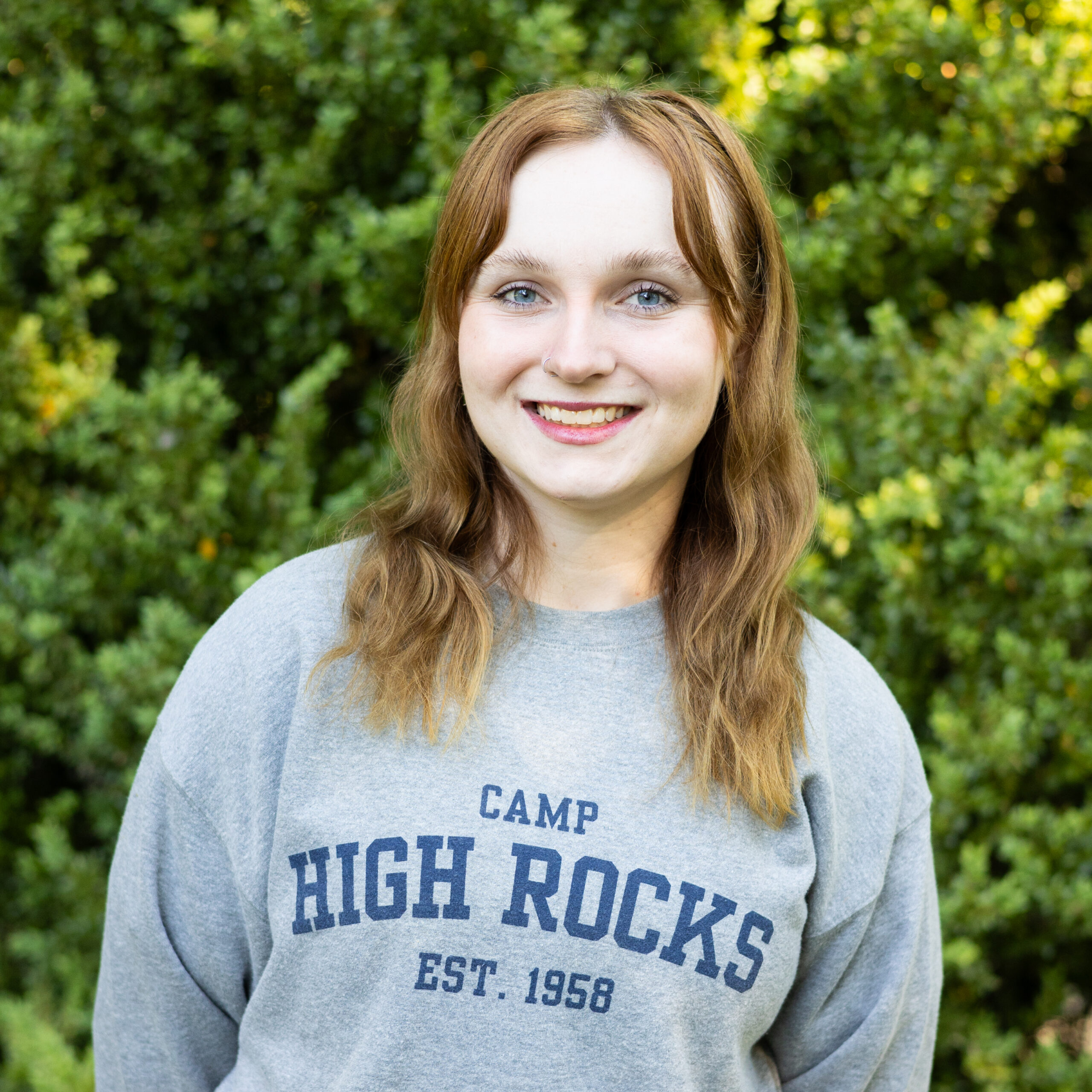 Smiling woman with long brown hair outdoors, wearing a gray sweatshirt with blue text, green foliage in the background.