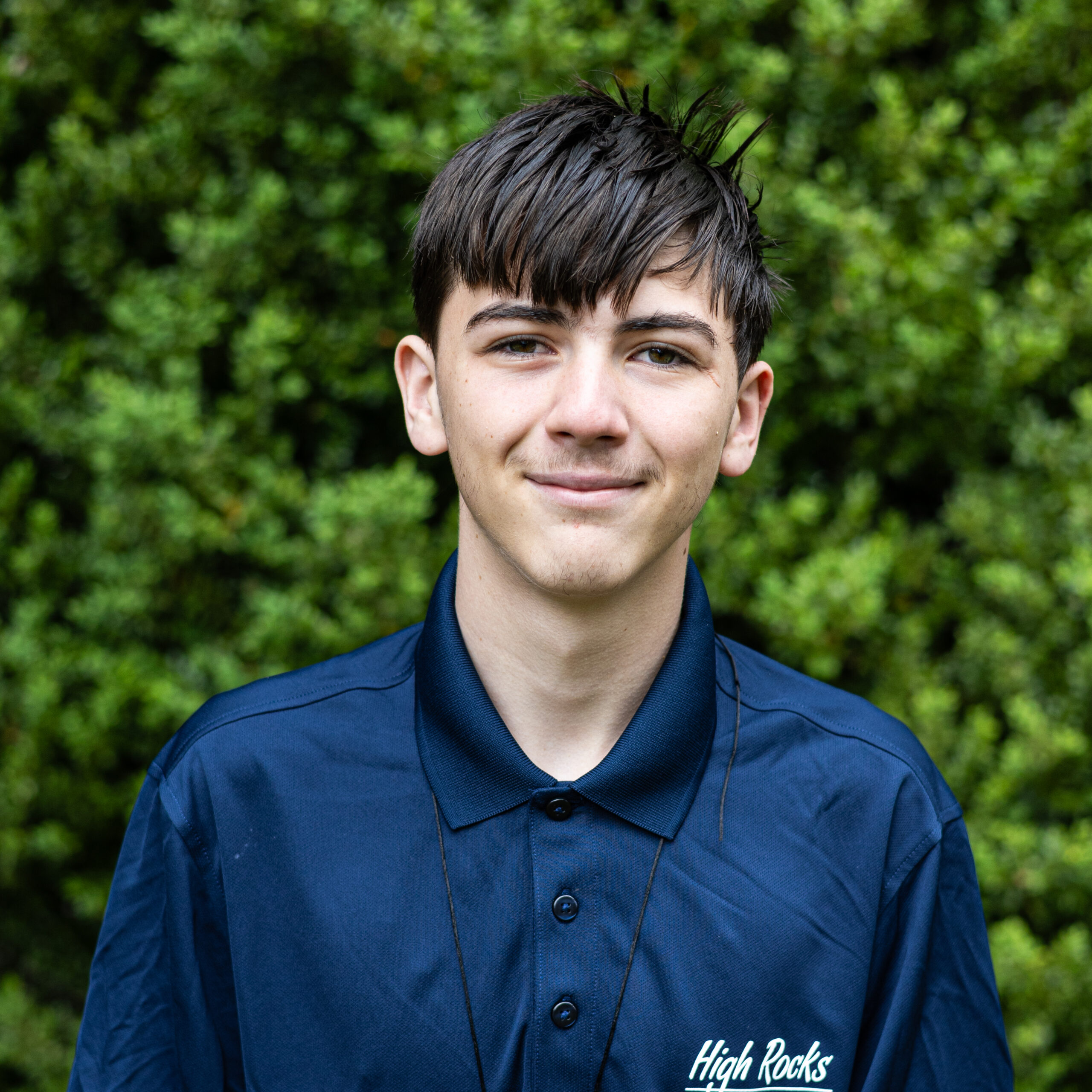 Close-up portrait of a young man with dark, wet-looking hair wearing a blue shirt, greenery behind him; shirt shows 'High Rocks' logo.