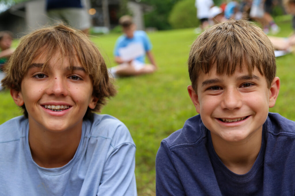 Two young boys smile at the camera in a park, their faces close together with a green field behind them.