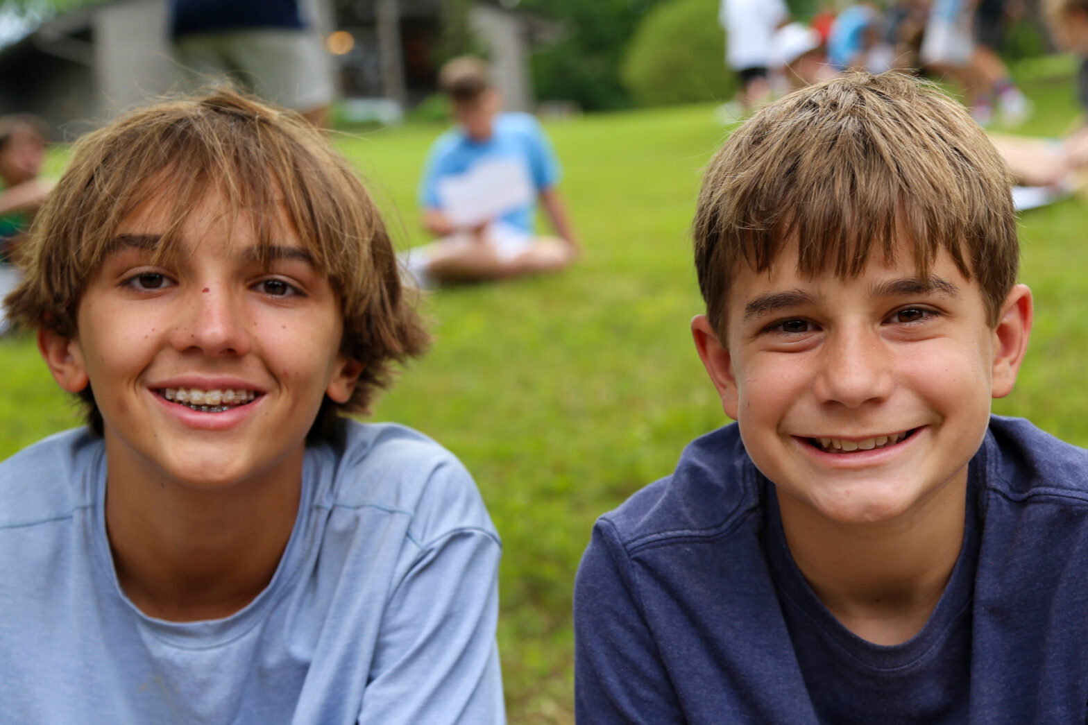 Two young boys smile at the camera in a park, their faces close together with a green field behind them.