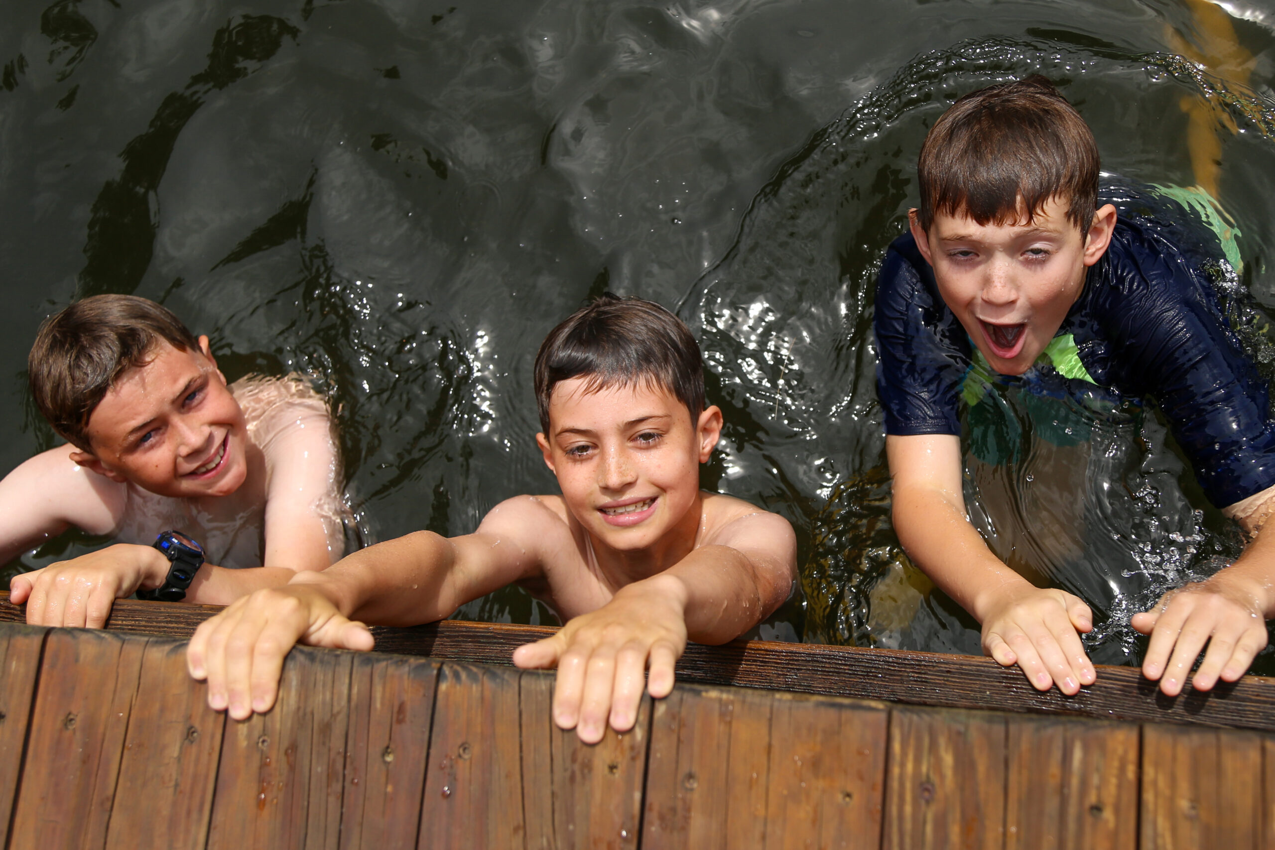 Three boys lean over a wooden railing into dark water, smiling as they splash around together.
