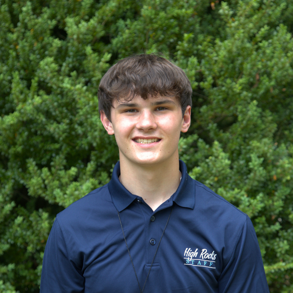 Smiling young man in a navy polo with High Rocks STAFF logo, posing in front of green bushes outdoors.