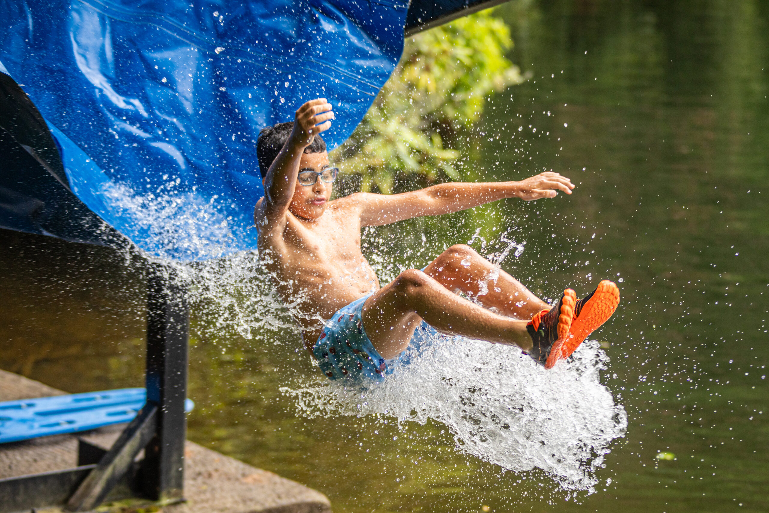 Boy in swim trunks jumps off a blue water slide into a lake, water splashing around him.
