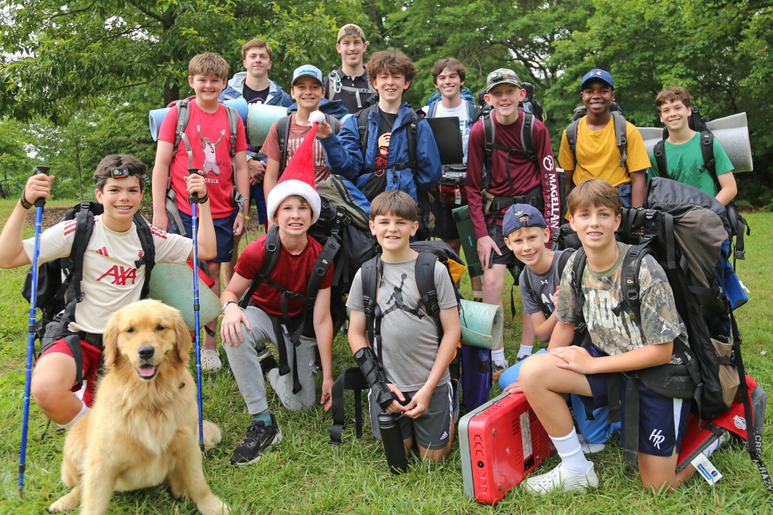 Group of smiling outdoor club members with backpacks posing for a photo in a grassy park, a golden retriever sits at the front left wearing a headlamp, and camping gear rests on their shoulders.