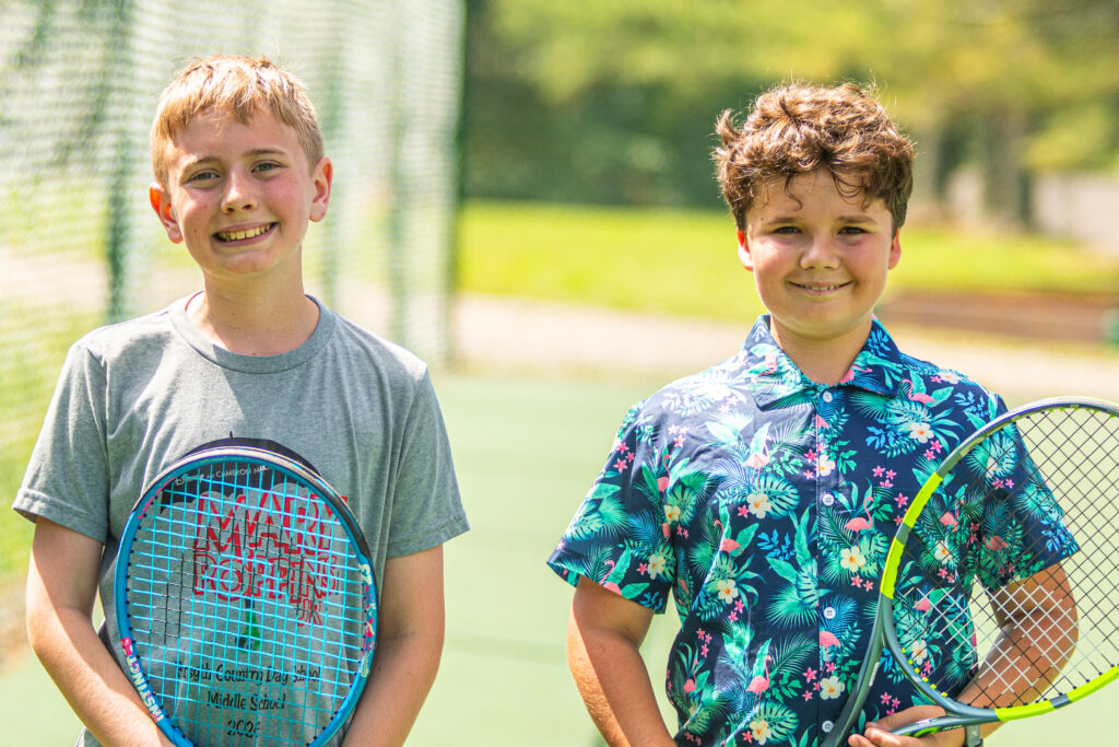 Two boys posing with tennis rackets on a sunny outdoor court, smiling at the camera.