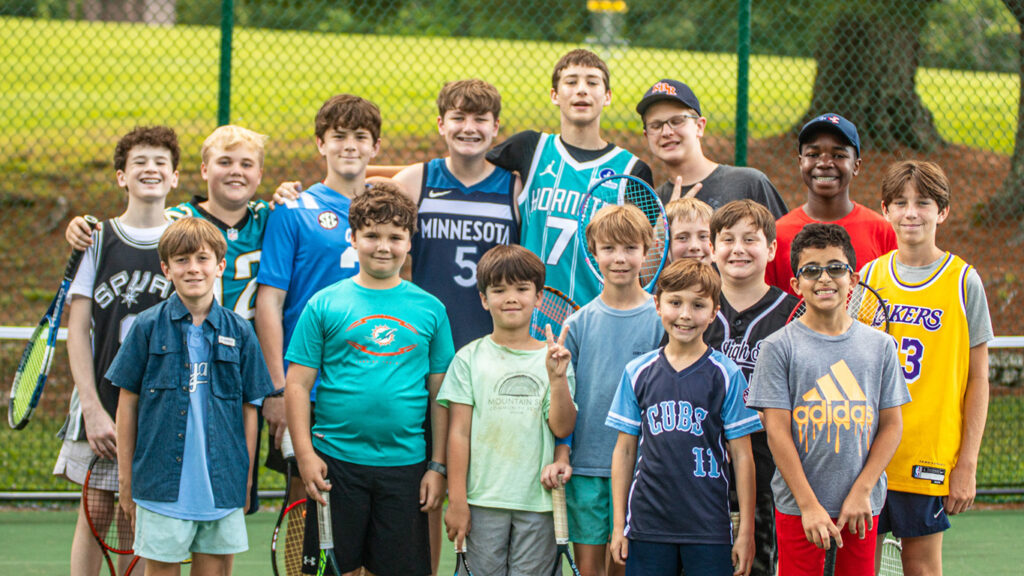 Group of kids on a tennis court posing for a team photo, holding rackets and smiling.