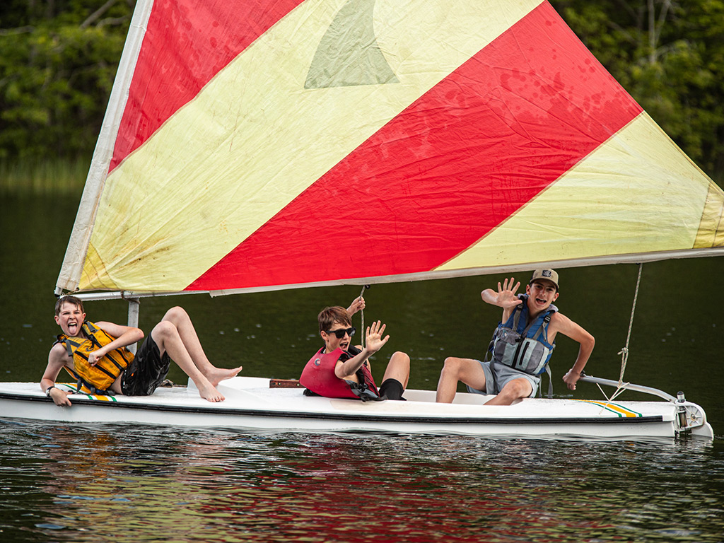 Three children on a small sailboat with a large colorful sail on a lake, waving and smiling.