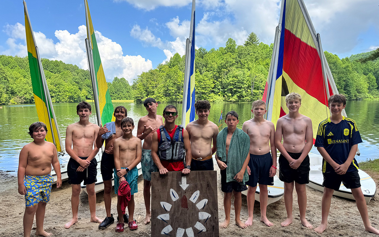 Group of boys at a lake camp posing with sailboats and colorful masts behind them on the shore.