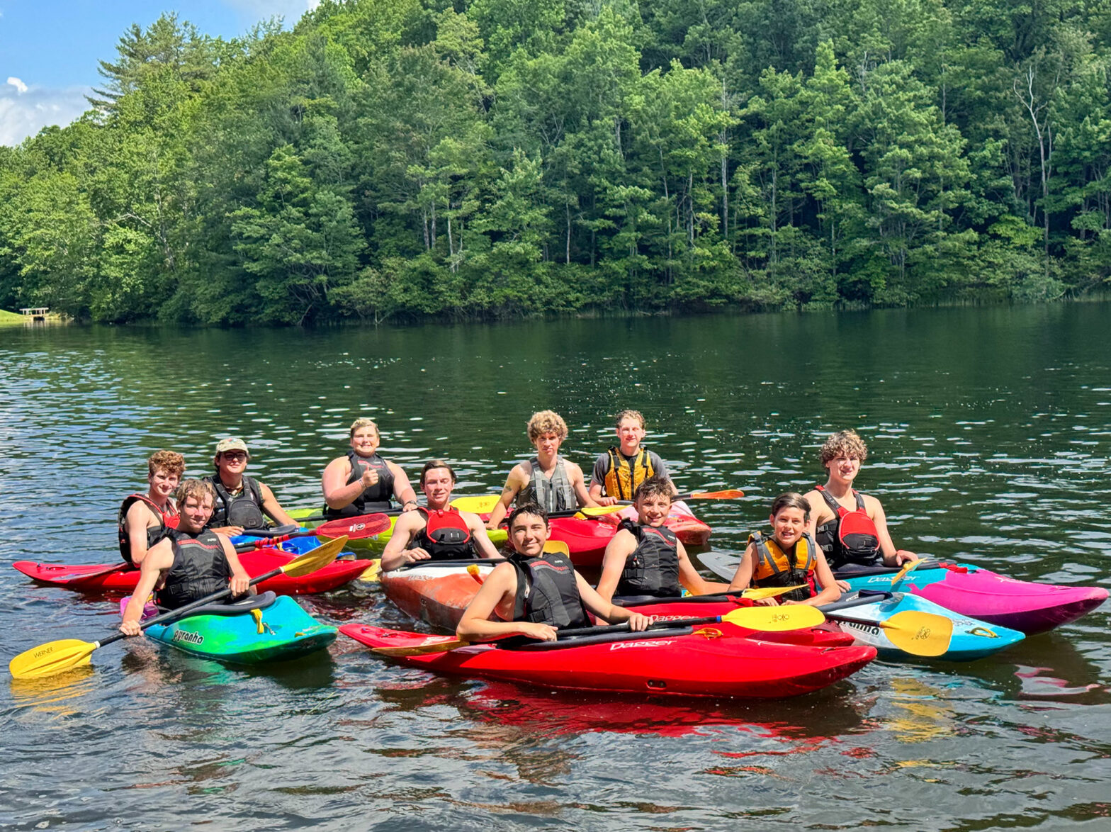 Group of people in colorful kayaks posing on a lake with a forested shoreline in the background.