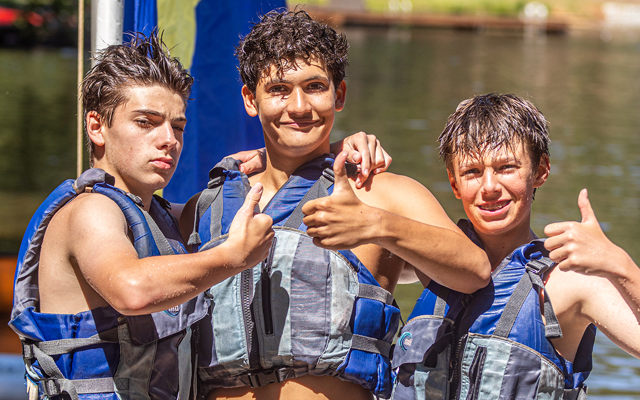 Three smiling teens in blue life jackets give thumbs up by a lake, celebrating after a water activity.