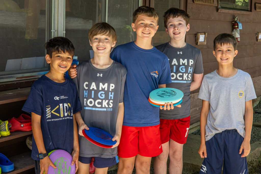 Five boys stand outdoors in front of a wooden building, smiling, each holding colorful frisbees ready to play. (Camp or outdoor activity scene)