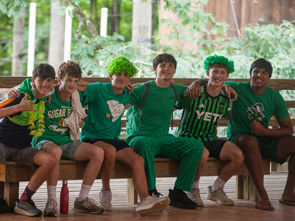 Six young men in green shirts sit arm-in-arm on a wooden bench outdoors, smiling under a leafy backdrop with camp vibes.