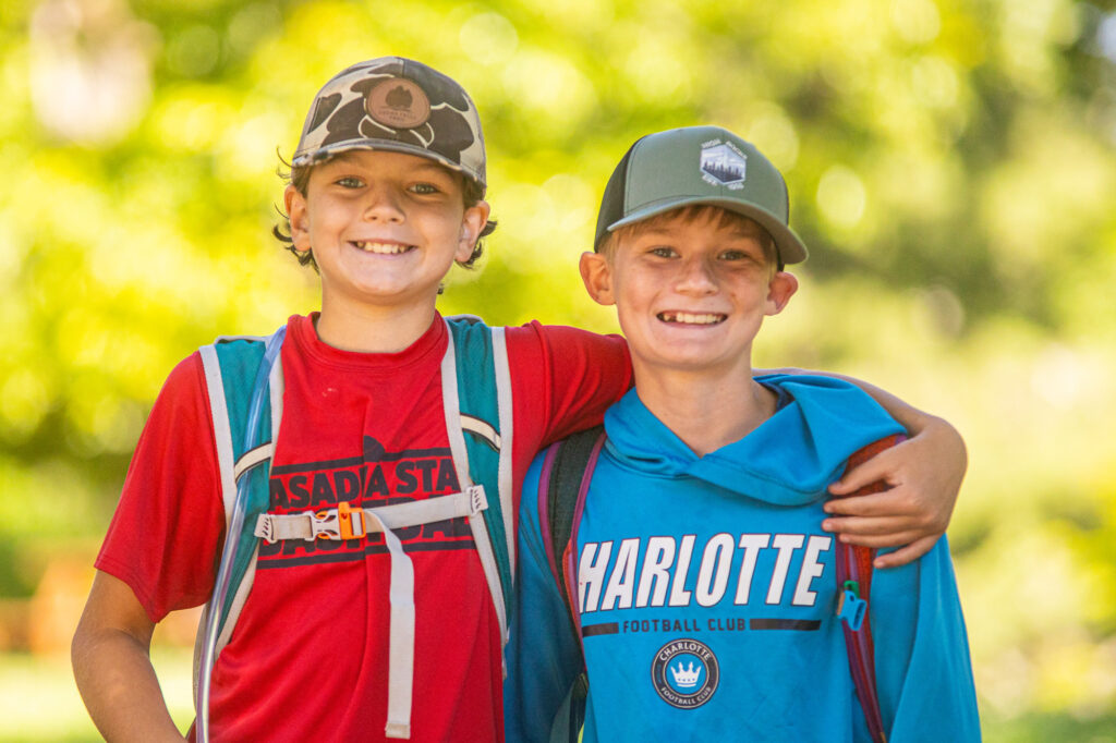 Two smiling boys with backpacks pose outdoors, one in red shirt and camo hat, the other in blue Charlotte FC shirt, arms around each other.