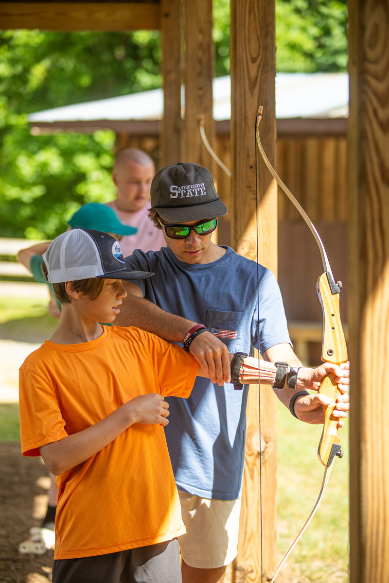 Boy in orange shirt receives archery instruction from a man in a blue shirt at an outdoor wooden shooting station, bow in hand.